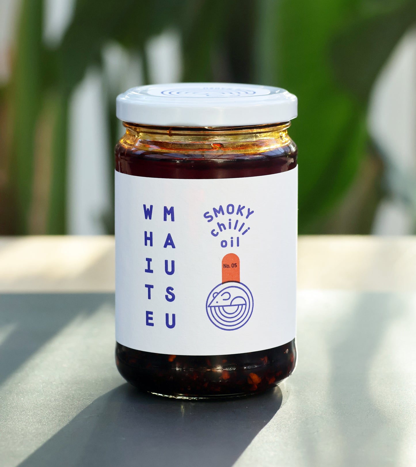 Ein 100-ml-Glas White Mausu | Rauchiges Chiliöl von White Mausu Ltd. ruht auf einer grauen Oberfläche, mit verschwommenen grünen Pflanzen im Hintergrund, die den rauchigen Geschmack hervorheben.