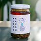 Ein 460-g-Glasgefäß mit Erdnuss Rayu Extra Hot von White Mausu Ltd. steht auf einem Tisch. Das weiße Etikett und der Deckel kennzeichnen das extra scharfe Chili-Öl mit sichtbaren Sesamsamen vor einem verschwommenen grünen Hintergrund.