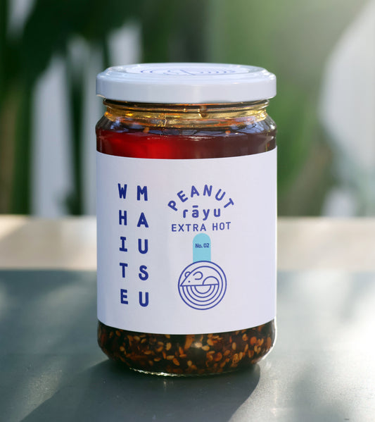 Ein 460-g-Glasgefäß mit Erdnuss Rayu Extra Hot von White Mausu Ltd. steht auf einem Tisch. Das weiße Etikett und der Deckel kennzeichnen das extra scharfe Chili-Öl mit sichtbaren Sesamsamen vor einem verschwommenen grünen Hintergrund.