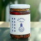 Ein 440-g-Glas der White Mausu | Erdnuss Rayu Sauce von White Mausu Ltd. mit blauem Etikett steht auf einer Oberfläche mit verschwommenem grünem Hintergrund und verströmt eine verlockende Umami-Atmosphäre.