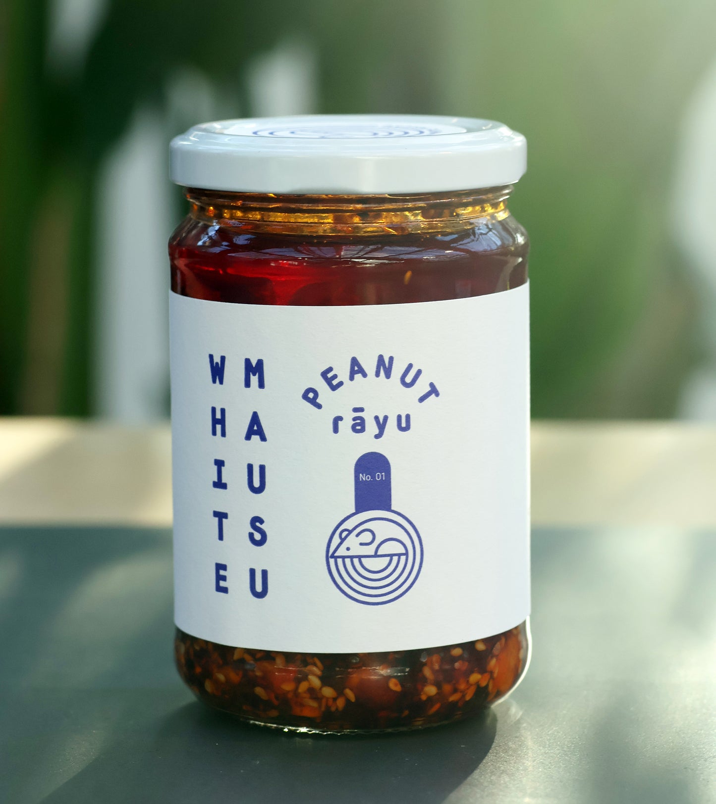 Ein 440-g-Glas der White Mausu | Erdnuss Rayu Sauce von White Mausu Ltd. mit blauem Etikett steht auf einer Oberfläche mit verschwommenem grünem Hintergrund und verströmt eine verlockende Umami-Atmosphäre.