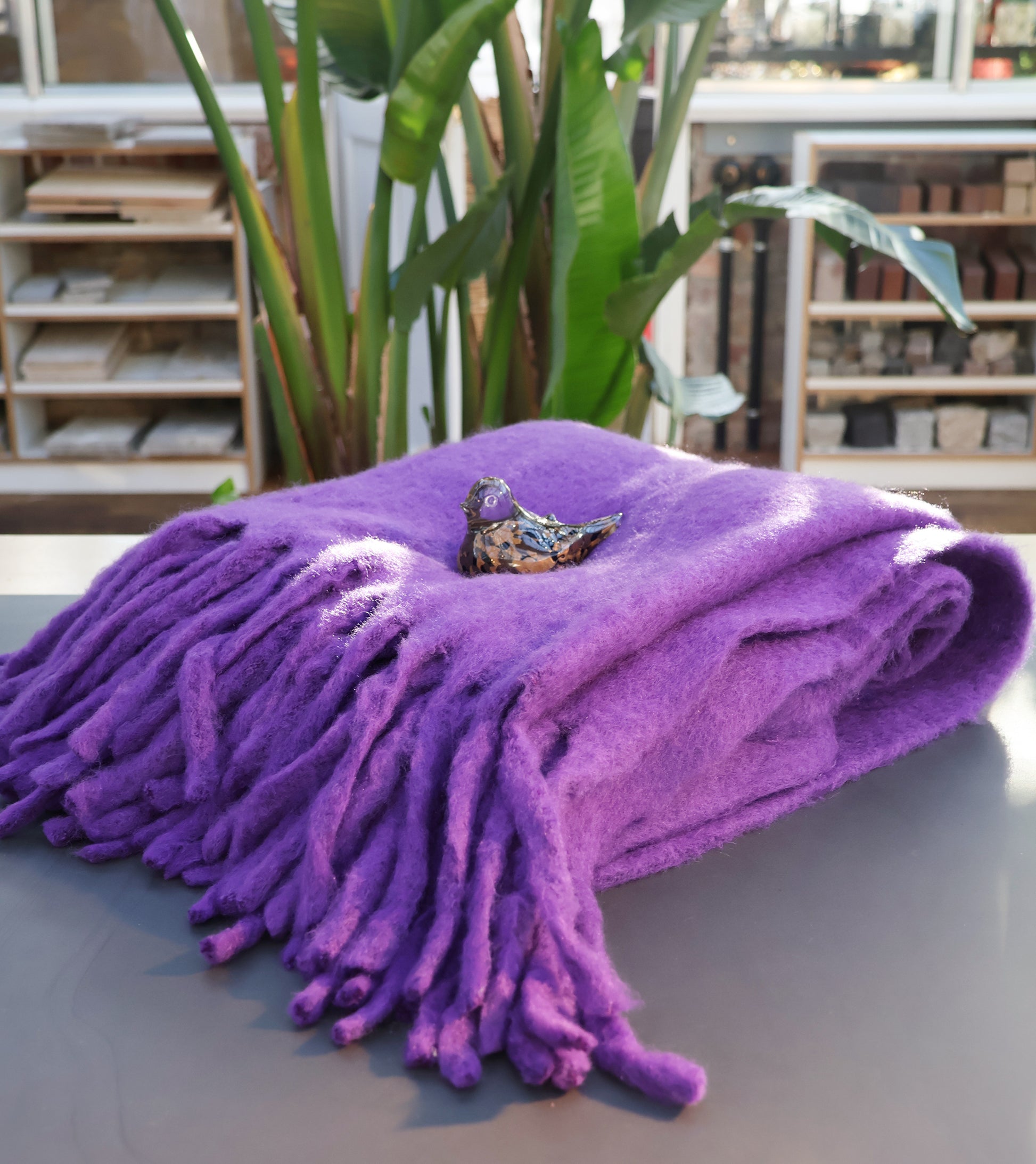 Eine gefaltete Werner Voss | Lila Decke aus Wollmischung liegt auf einem Tisch, auf dem eine kleine Vogelfigur aus Keramik steht; im Hintergrund stehen Grünpflanzen und Regale.