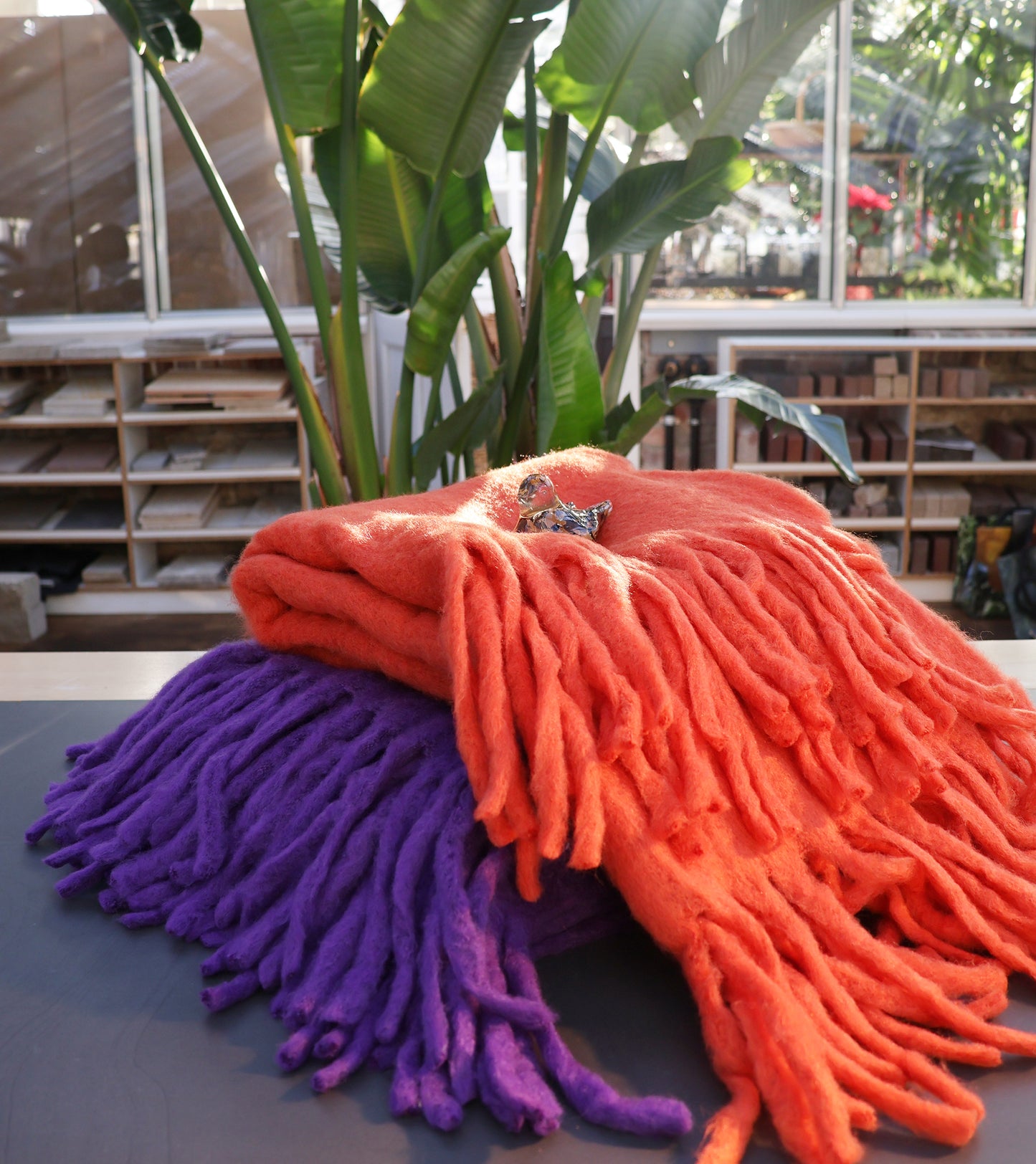 Die orangefarbene Decke aus Wollmischung von Werner Voss liegt gefaltet auf dem Tisch im Sonnenlicht. Im Hintergrund stehen eine große grüne Pflanze und Regale mit stilvollen Werner Voss-Stücken.