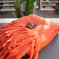 Eine kleine Vogelfigur aus Keramik sitzt auf der Werner Voss | Orange Decke aus Wollmischung, die gefaltet auf einem dunklen Tisch mit grünen Pflanzen und Regalen im Hintergrund steht.