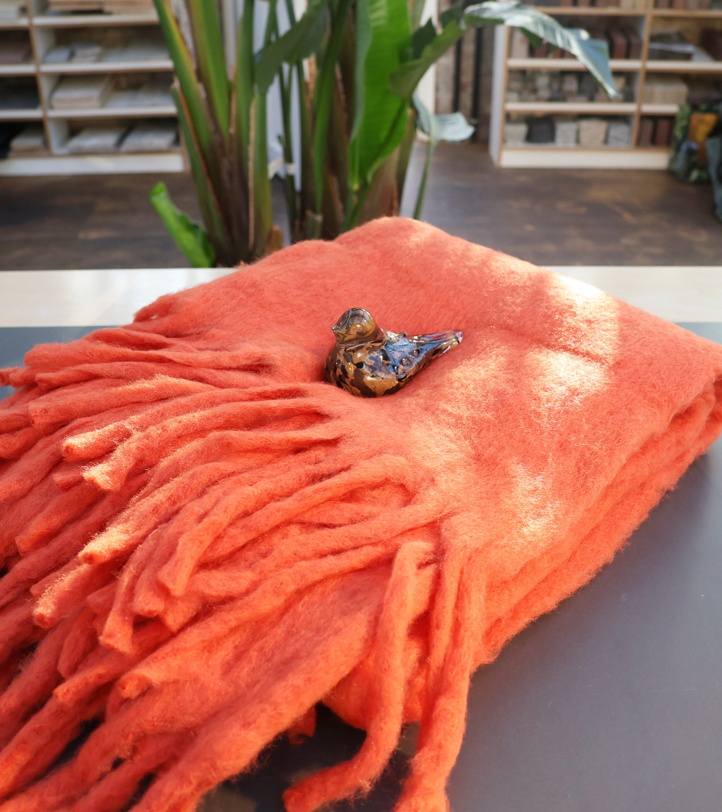 Eine kleine Vogelfigur aus Keramik sitzt auf der Werner Voss | Orange Decke aus Wollmischung, die gefaltet auf einem dunklen Tisch mit grünen Pflanzen und Regalen im Hintergrund steht.