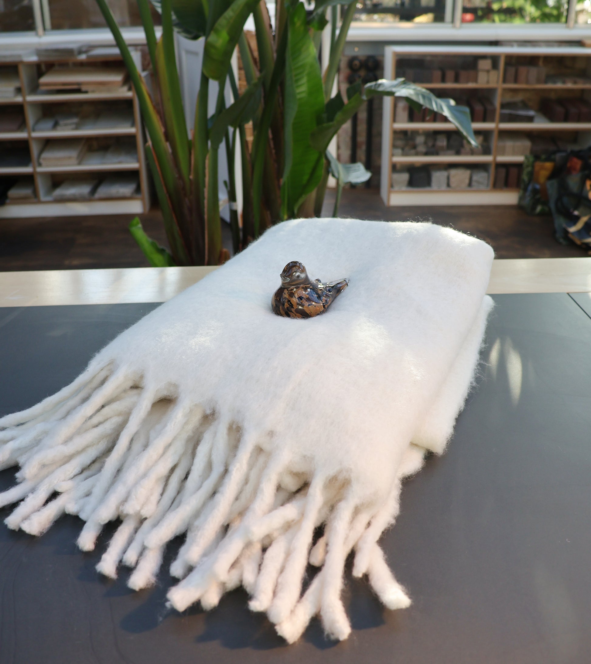 Eine ordentlich gefaltete, weiße Decke aus Wollmischung von Werner Voss liegt auf einem Tisch, darauf ein kleiner dekorativer Keramikvogel. Im Hintergrund sind grüne Pflanzen und Regale mit Büchern.