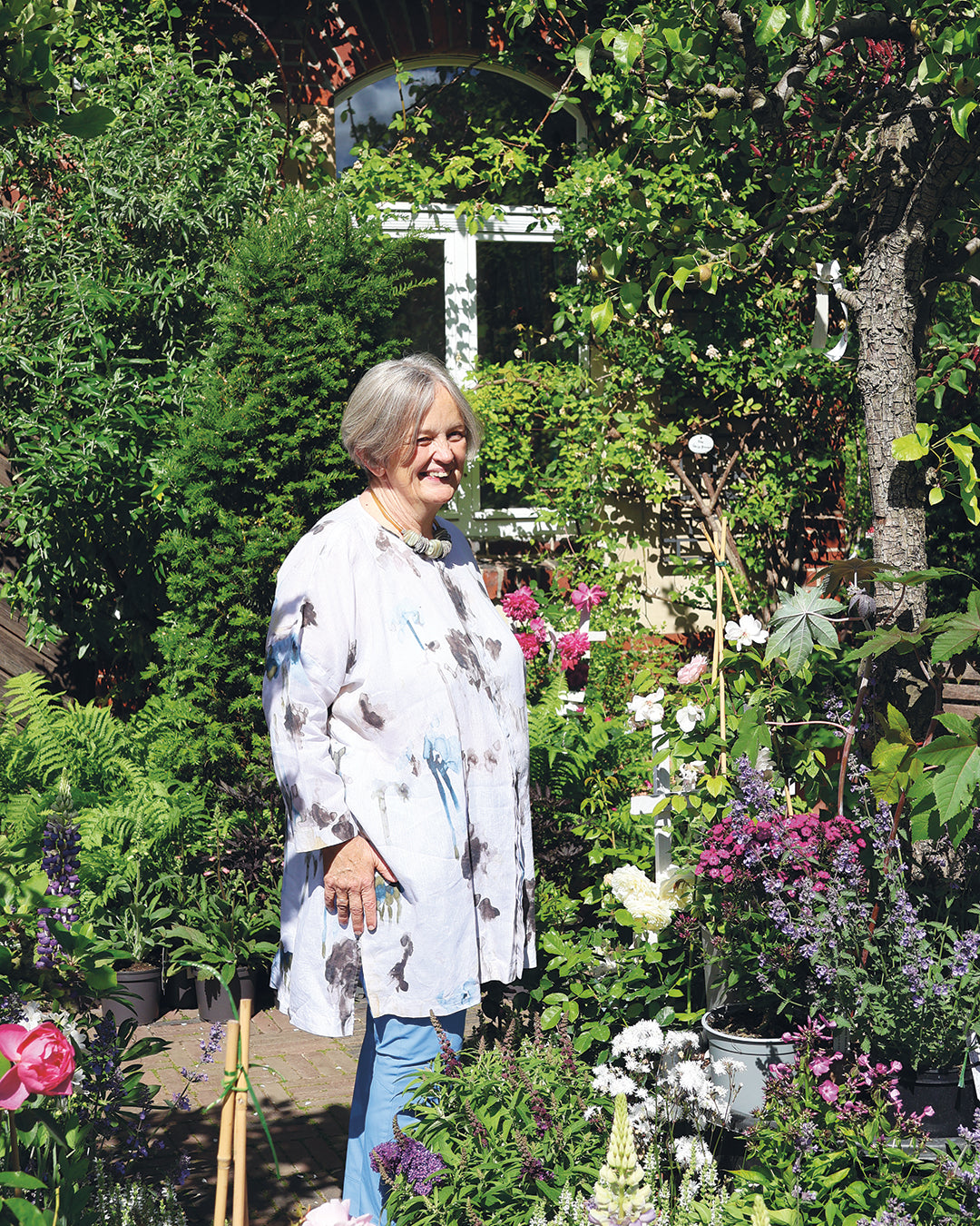Eine Frau steht in einem üppigen Garten mit professioneller Beetgestaltung inmitten von Topfblumen und Grünpflanzen für Mi., den 10.3. - Do., 11.3.2027 | 10.00 – 16.30 Uhr ZWEITÄGIGER MEISTERKURS Gestalten Sie Ihr eigenes Beet.