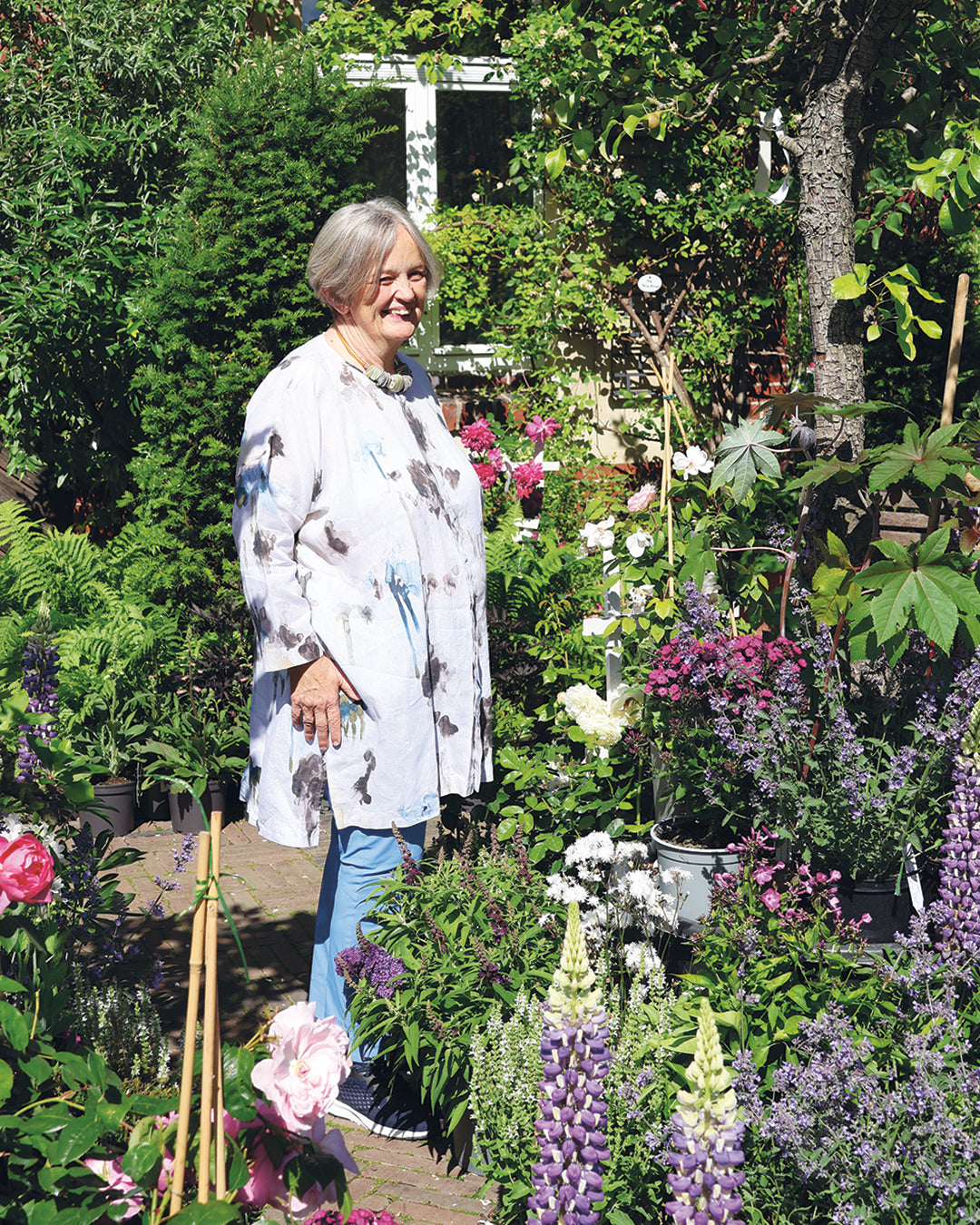 Eine ältere Frau lächelt in einem lebendigen Garten und bewundert ihr Blumenbeet, das sie beim "ZWEI-TAGES-MASTERCLASS Gestalten Sie Ihr eigenes Beet" der Königlichen Gartenakademie (Mi. 28.10 - Do. 29.10.2026 | 10:00-16:30) angelegt hat. Das Sonnenlicht erhellt die fröhliche Szene.