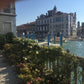 Ein sonniger Blick auf die Kanäle und historischen Gebäude Venedigs, mit blühenden Blumen und einem Metallzaun in den Gärten in Venedig. In der Ecke ist ein Finger zu sehen. Ereignis: Di. 03.11.2026 | 14:30-16:00 AFTERNOON TEA by Königliche Gartenakademie.