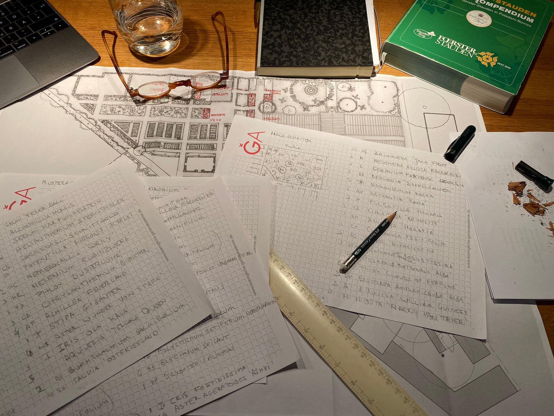 Ein vollgestopfter Schreibtisch mit Notizen, Zeichnungen und einem grünen Buch bildet die Kulisse - ideal für die Planung der "ZWEI-TAGES-MASTERCLASS Gestalten Sie Ihr eigenes Beet" der Königlichen Gartenakademie am Mi. 28.10 - Do. 29.10.2026, 10:00-16:30.