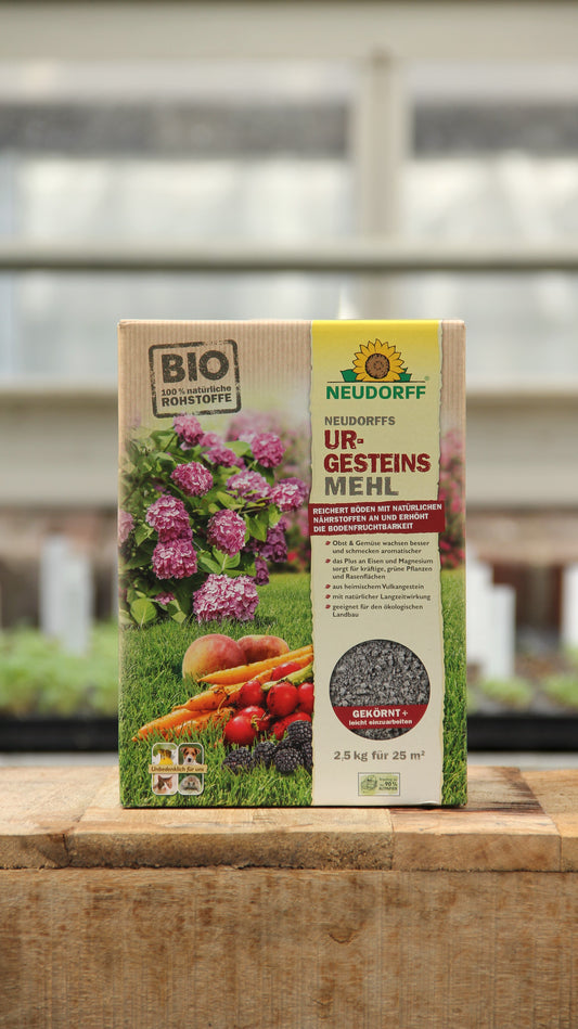 Eine Schachtel Neudorff Urgesteinsmehl aus Diabasgesteinsmehl vulkanischen Ursprungs (2,5 kg) steht auf einer Holzfläche, die Verpackung ist mit Gemüse und Blumen verziert. Ein Gewächshaus und Pflanzen sind sanft verschwommen im Hintergrund zu sehen.