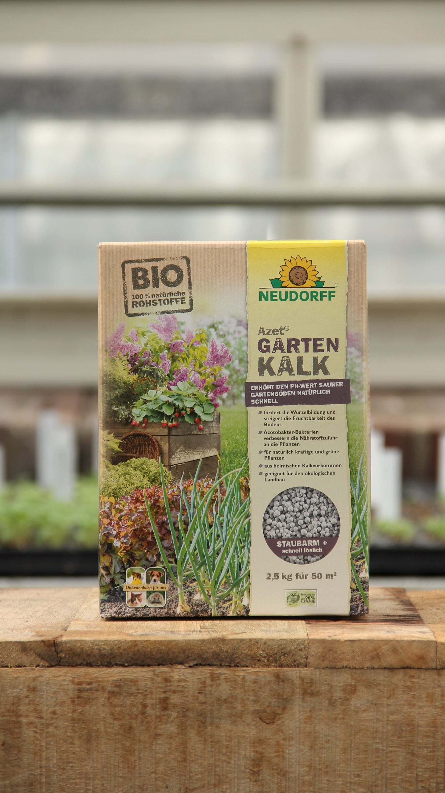 Eine 2,5 kg Kiste Neudorff | Gartenkalk Bodenverbesserer aus natürlichen Rohstoffen steht auf Holz, umrahmt von Grünpflanzen und einem unscharfen Gewächshaus. Die Verpackung zeigt die Verwendung zur Regulierung des pH-Werts im Boden, mit Informationen auf Deutsch.
