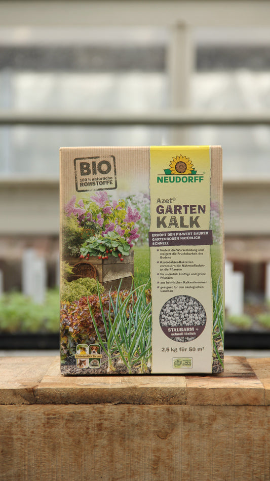 Eine 2,5 kg Kiste Neudorff | Gartenkalk Bodenverbesserer aus natürlichen Rohstoffen steht auf Holz, umrahmt von Grünpflanzen und einem unscharfen Gewächshaus. Die Verpackung zeigt die Verwendung zur Regulierung des pH-Werts im Boden, mit Informationen auf Deutsch.