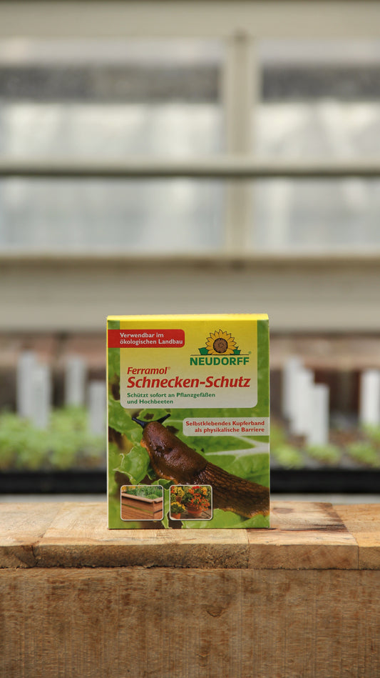Eine Schachtel mit Neudorff | Ferramol Schnecken-Schutz zur natürlichen Schneckenbekämpfung steht auf einer Holzfläche mit unscharfen Garten-Setzlingsschalen und einem Fenster im Hintergrund.