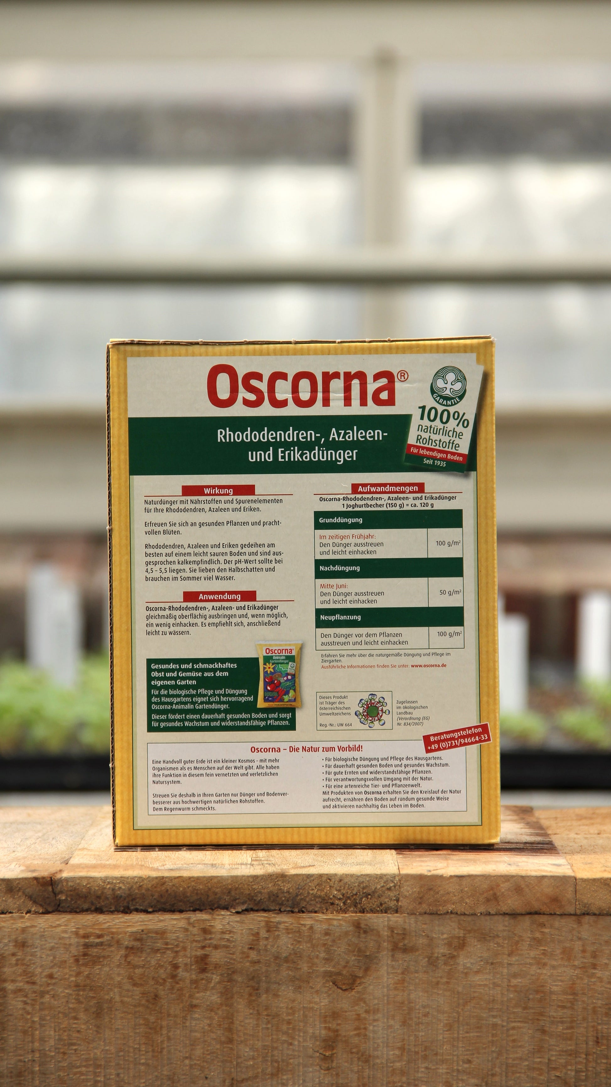 Eine 1 kg Kiste Oscorna Rhododendren-, Azaleen- und Erikadünger Naturdünger von Oscorna steht auf einer hölzernen Unterlage, ihr grün-rotes deutsches Etikett mit einer kleinen Abbildung vor einem Gewächshaushintergrund ist sichtbar.