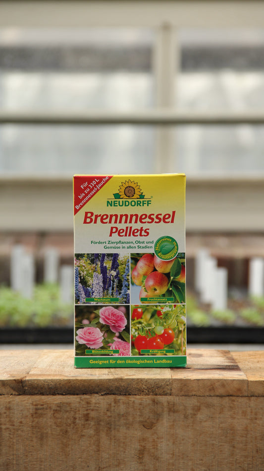 Eine Schachtel Neudorff | Brennnessel Pellets organischer Dünger 500 g mit Abbildungen von Rosen, Tomaten, Äpfeln und Kirschen steht auf einer Holzfläche mit einem unscharfen Gewächshaus im Hintergrund.