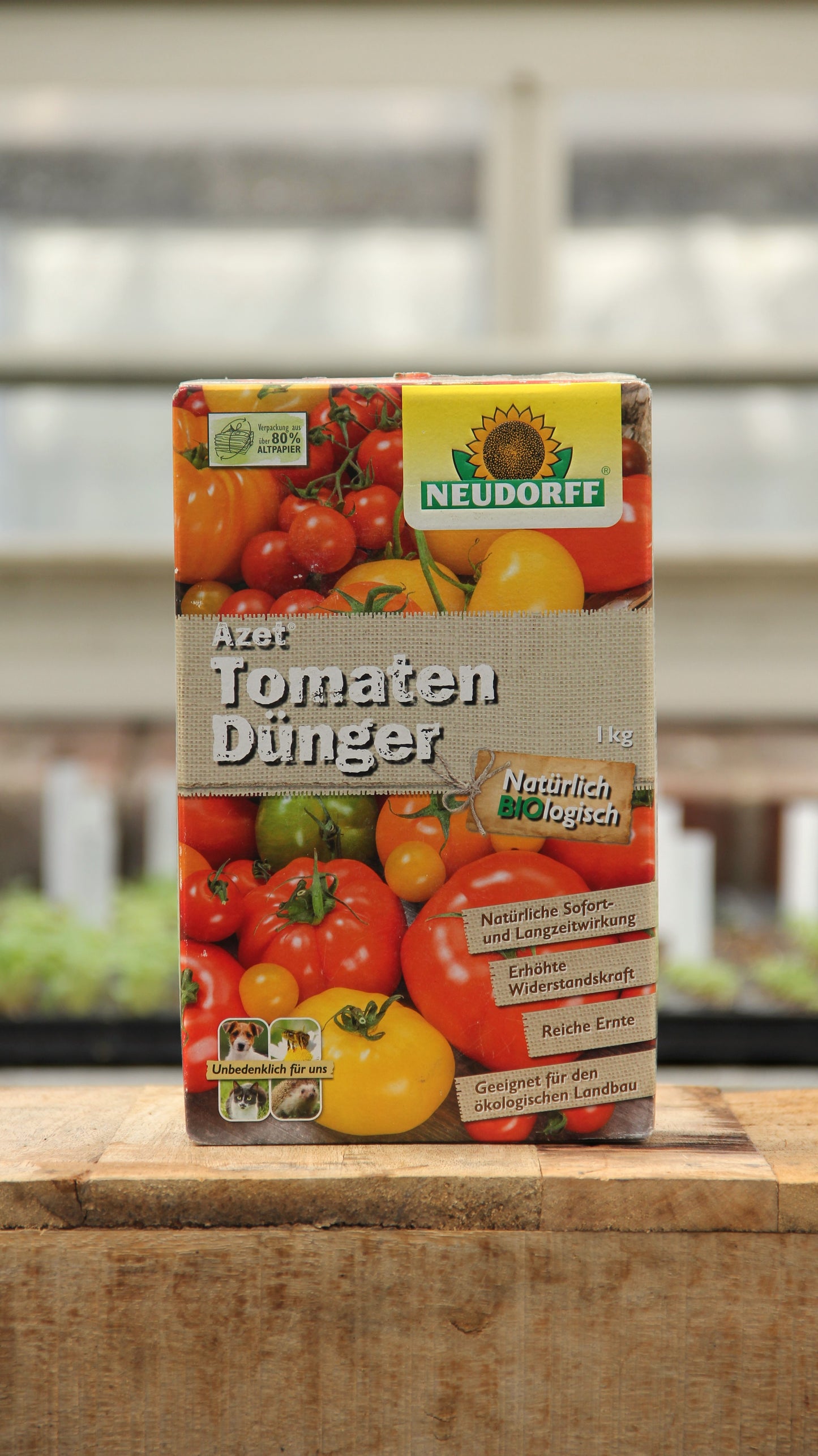 Auf einer hölzernen Unterlage steht ein 1 kg-Karton mit Tomatendünger von Neudorff Azet Tomaten Dünger, auf dem Bilder von roten und gelben Tomaten und ein deutscher Text abgebildet sind.
