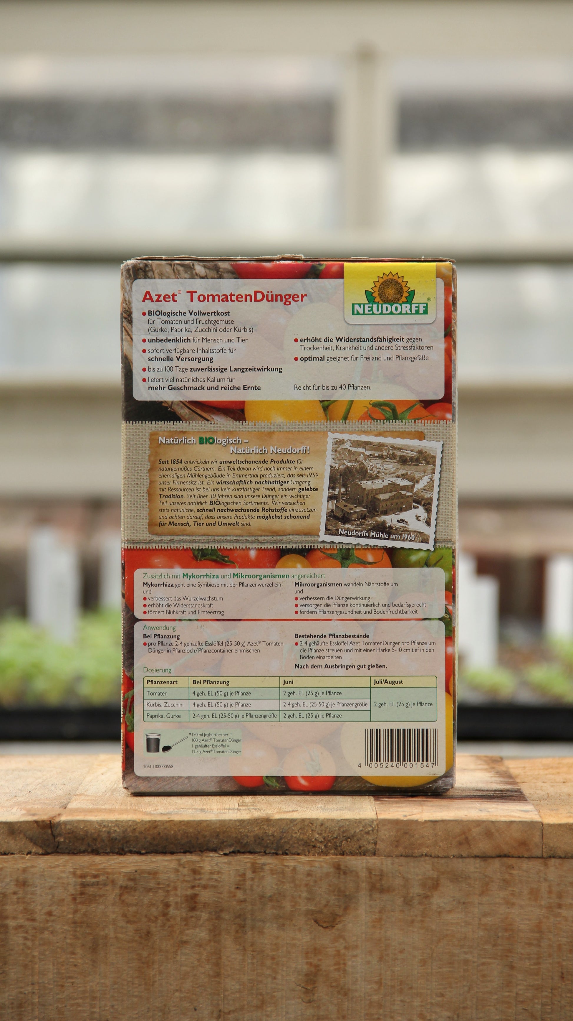 Eine 1 kg Kiste Neudorff | Azet Tomaten Dünger ist auf einer Holzfläche abgebildet, mit deutschen Produktangaben und Anweisungen auf der Rückseite. Im Hintergrund ist ein unscharfes Gewächshaus zu sehen.