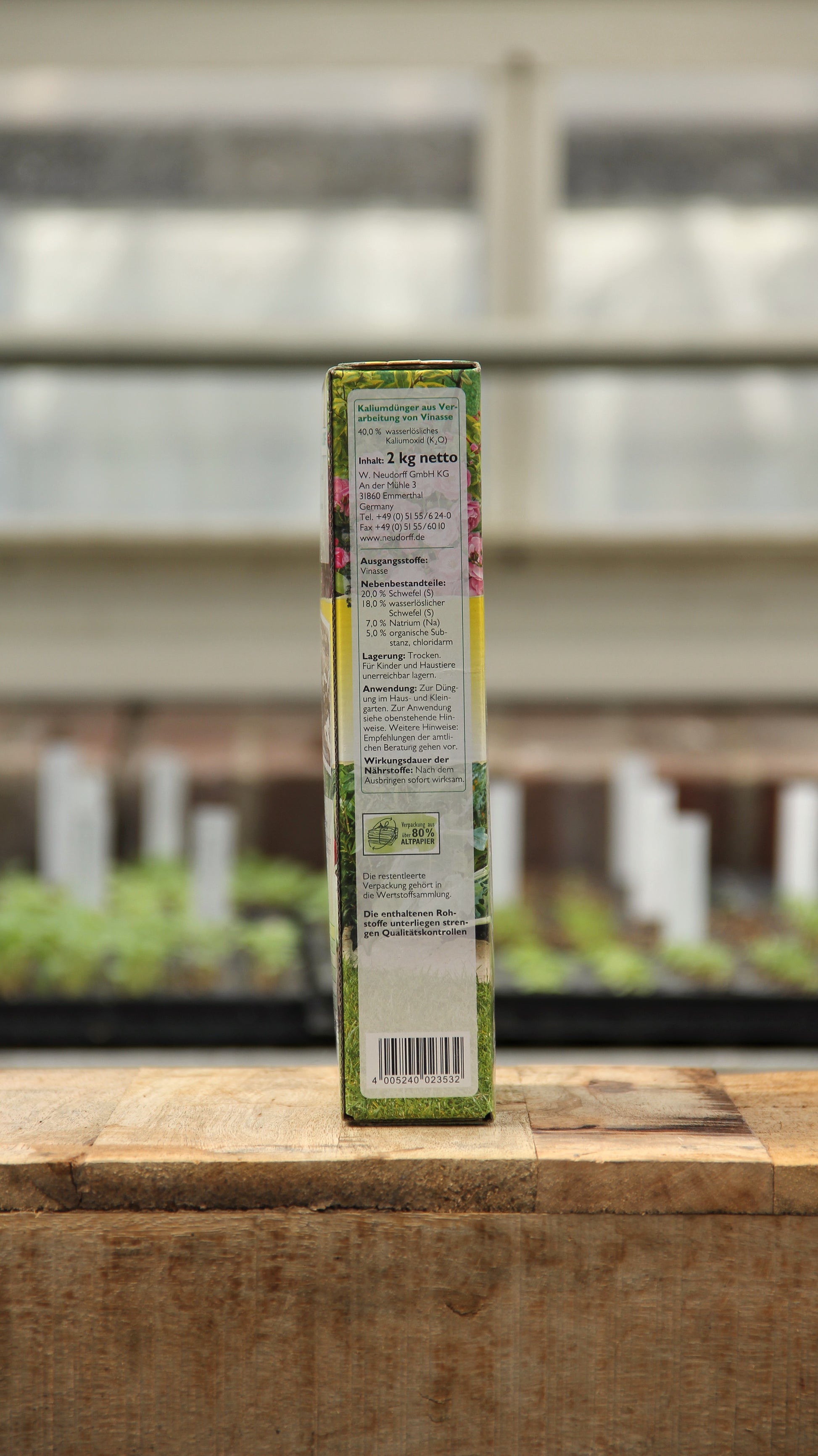 Eine rechteckige Schachtel Neudorff Azet Kalium Dünger 2 kg steht aufrecht auf einer hölzernen Oberfläche in einem Innenraum, mit verschwommenen Pflanzen und weißen Pflanzenetiketten im Hintergrund. Die Verpackung hebt den Kaliumdünger für frostbeständige Pflanzen hervor.