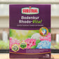 Ein 1 kg-Karton Substral | Bodenkur Rhodo-Vital für Hortensien, Azaleen und Kamelien steht auf einer Holzfläche und zeigt bunte Blumen auf der Verpackung, während im Hintergrund ein Gewächshaus zu sehen ist.