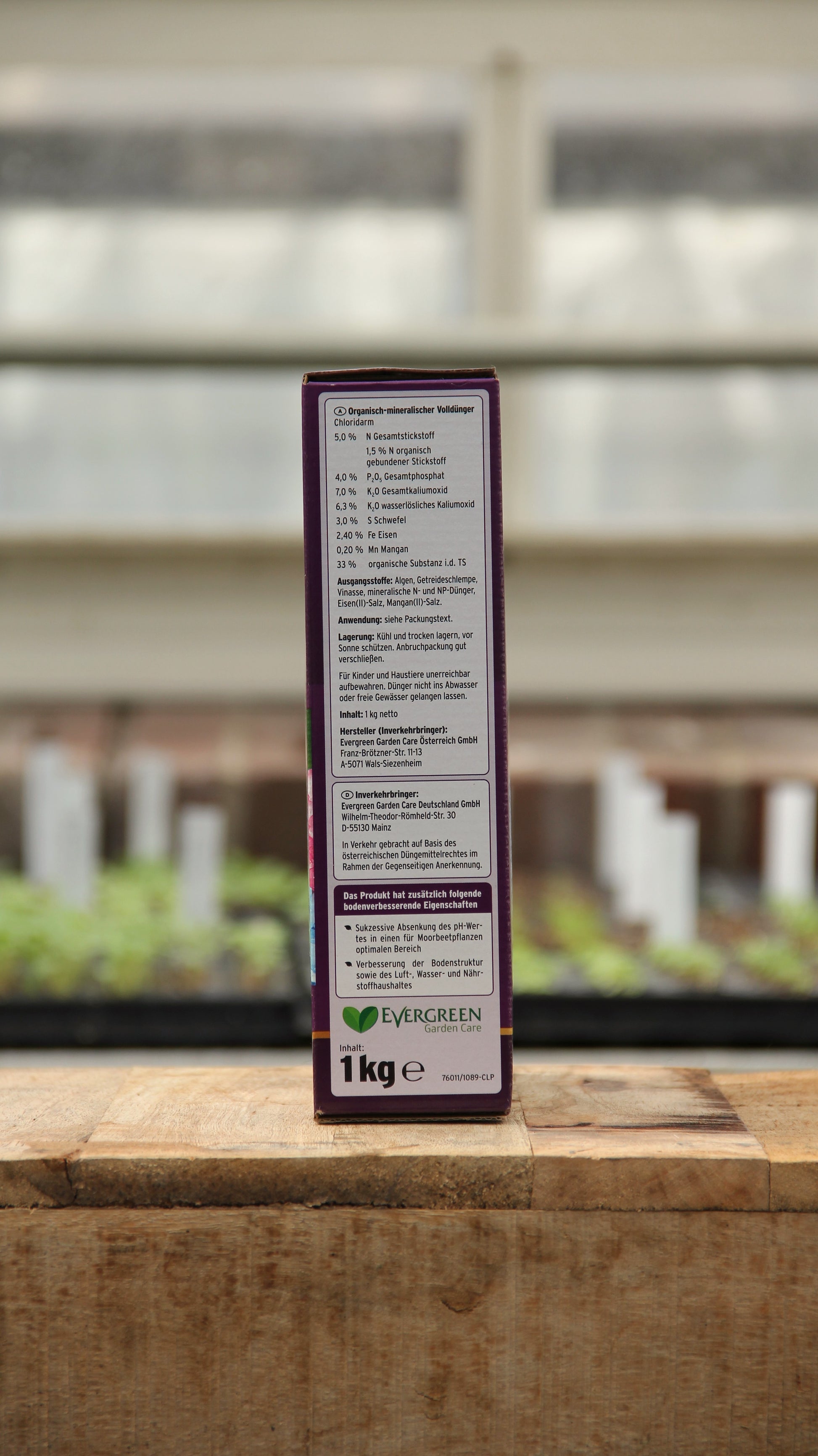 Eine rechteckige Schachtel Substral | Bodenkur Rhodo-Vital 1 kg steht aufrecht auf einer Holzfläche, mit Text und Anleitung, dahinter unscharfe Gewächshauspflanzen. Das Produkt hilft, den pH-Wert für ein gesundes Wachstum zu senken.