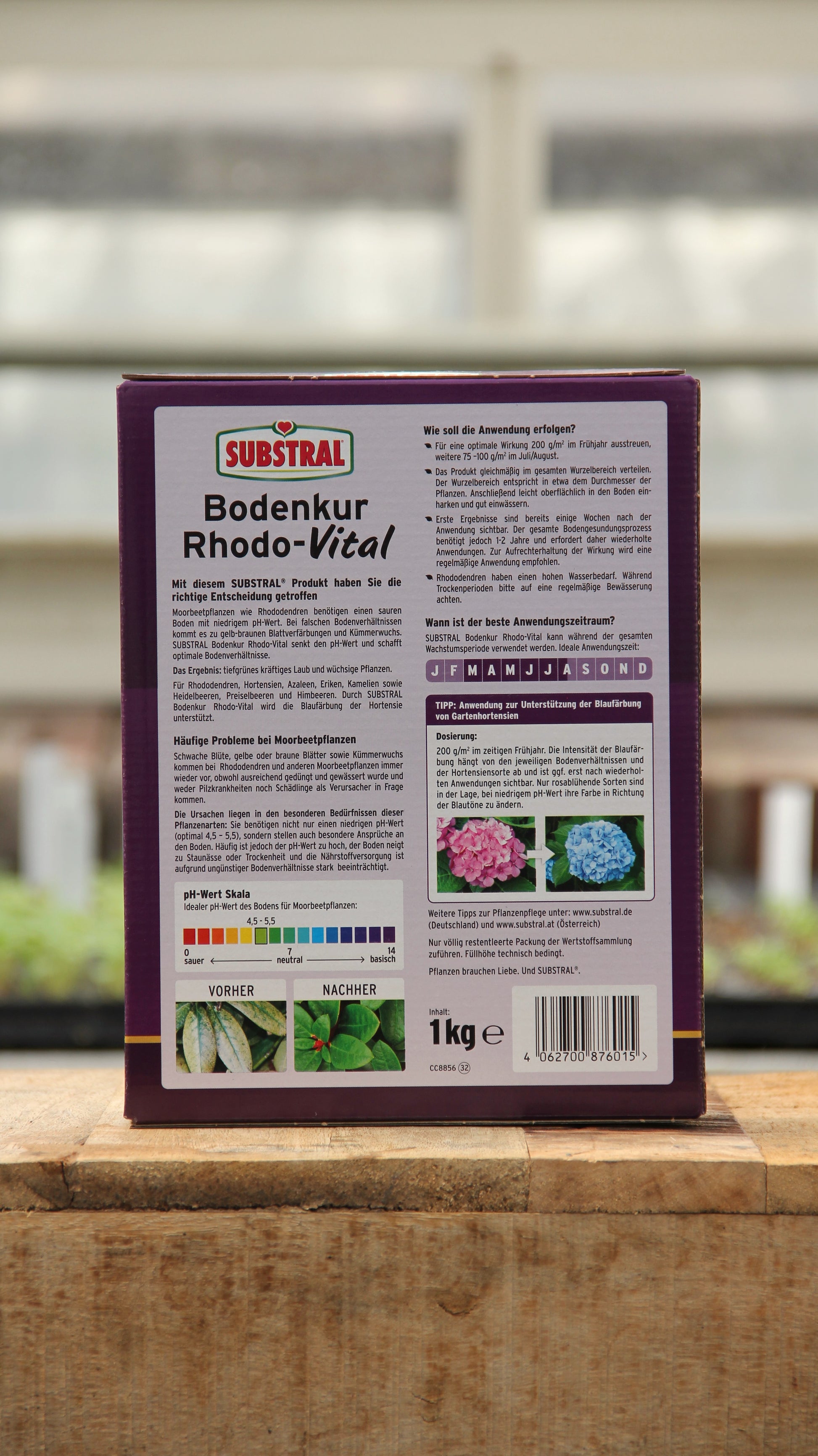 Ein 1 kg-Karton Substral Bodenkur Rhodo-Vital für Moorbeetpflanzen steht auf Holz, seine violette Verpackung enthält Anleitungen, Blumenbilder und eine Vorher-Nachher-Tabelle, die hilft, den pH-Wert zu senken.