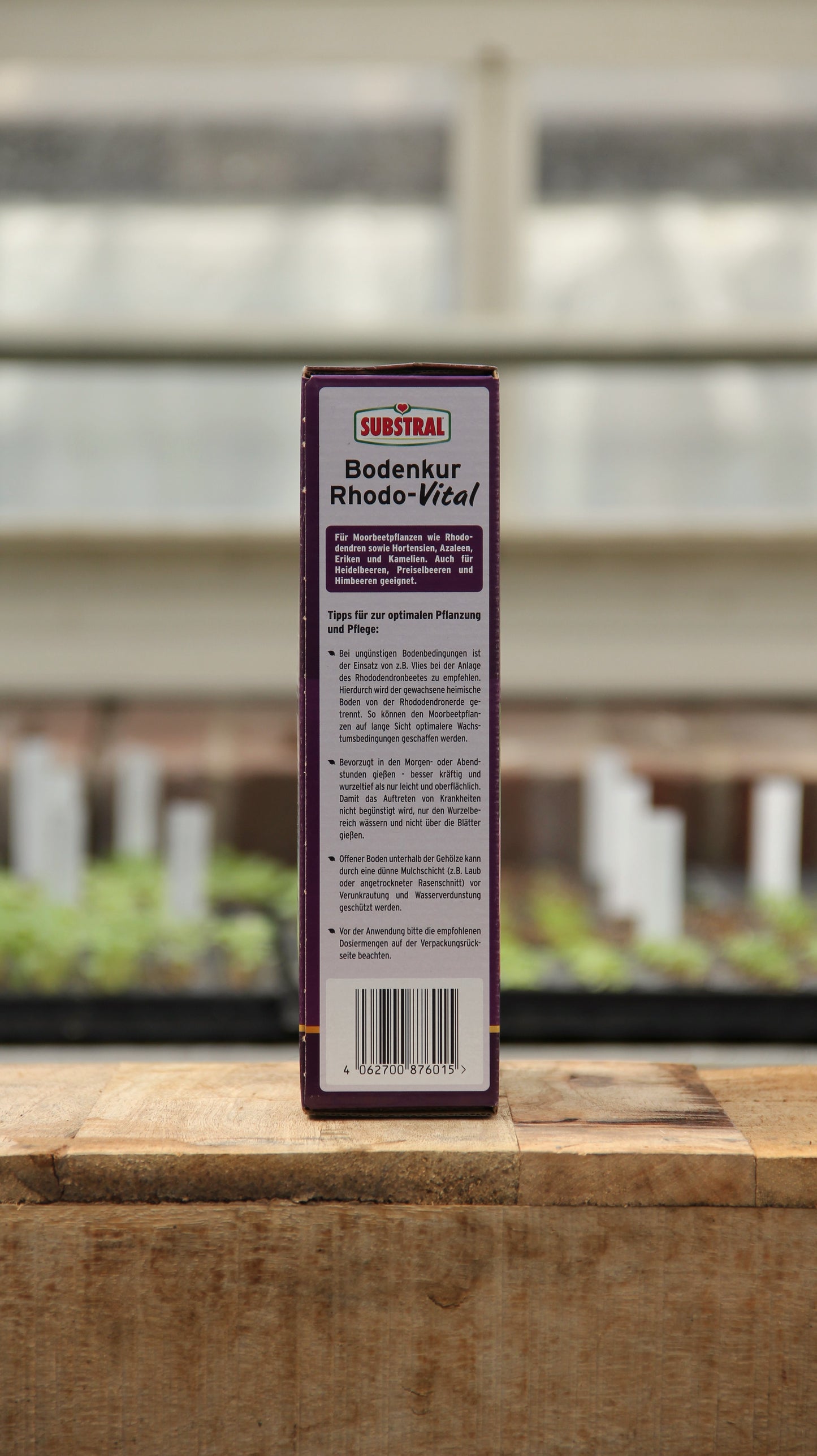 Eine rechteckige Schachtel Substral | Bodenkur Rhodo-Vital 1 kg steht auf einer hölzernen Unterlage und zeigt auf der Rückseite einen deutschen Text und eine Anleitung, während im Hintergrund Pflanzen und ein Fenster verschwimmen.