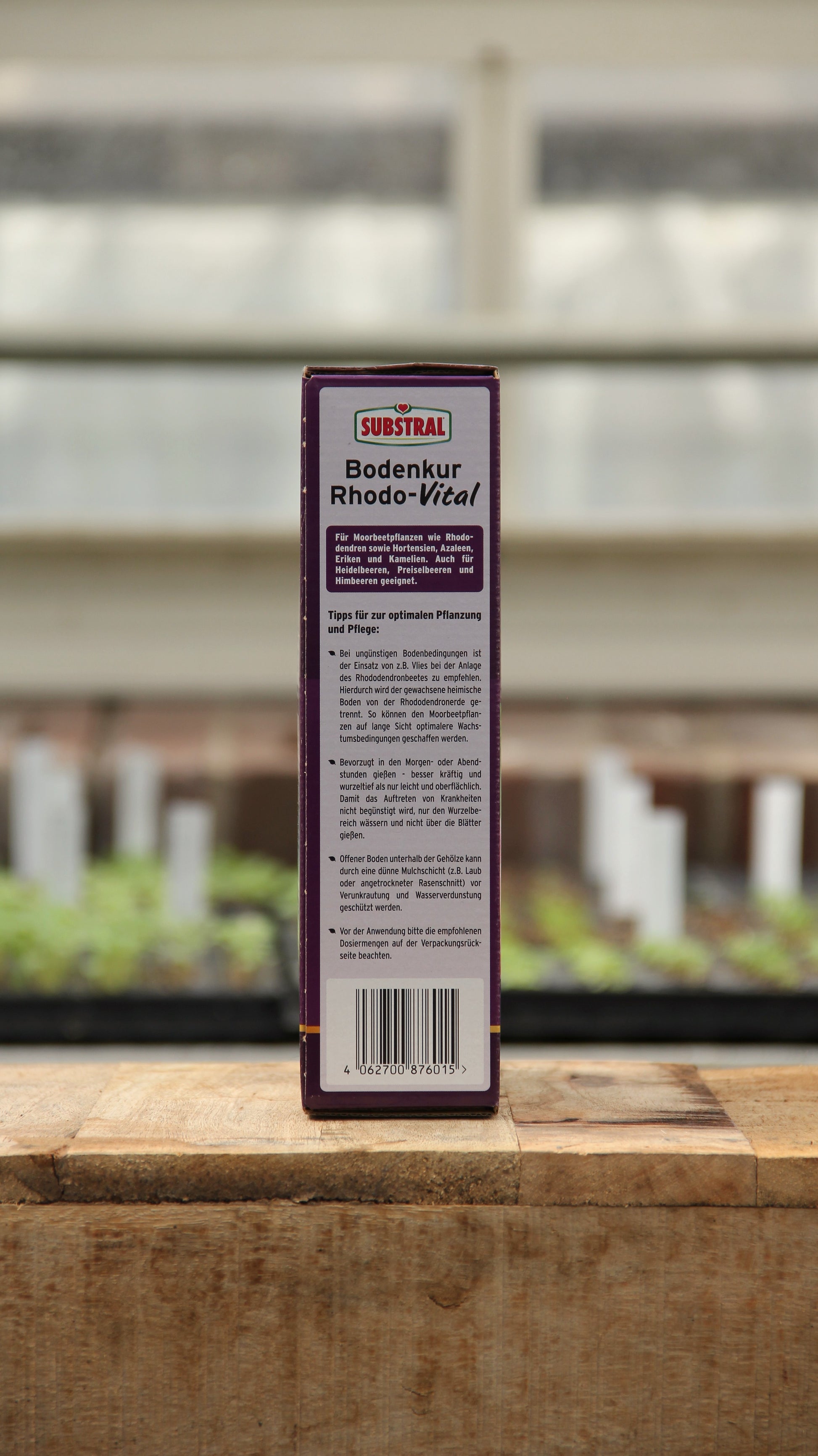 Eine rechteckige Schachtel Substral | Bodenkur Rhodo-Vital 1 kg steht auf einer hölzernen Unterlage und zeigt auf der Rückseite einen deutschen Text und eine Anleitung, während im Hintergrund Pflanzen und ein Fenster verschwimmen.