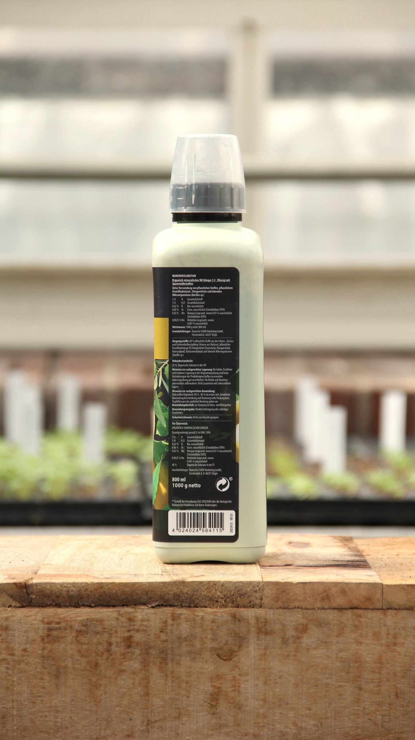 Eine Flasche Cuxin | Flüssigdünger Mediterrane Pflanzen & Zitruspflanzen Bio 800 ml mit schwarzem Etikett und klarem Verschluss steht auf Holz. Das Etikett zeigt Text, grüne Blätter, und im Hintergrund sind unscharfe Pflanzen mit weißen Pflanzenmarkern.