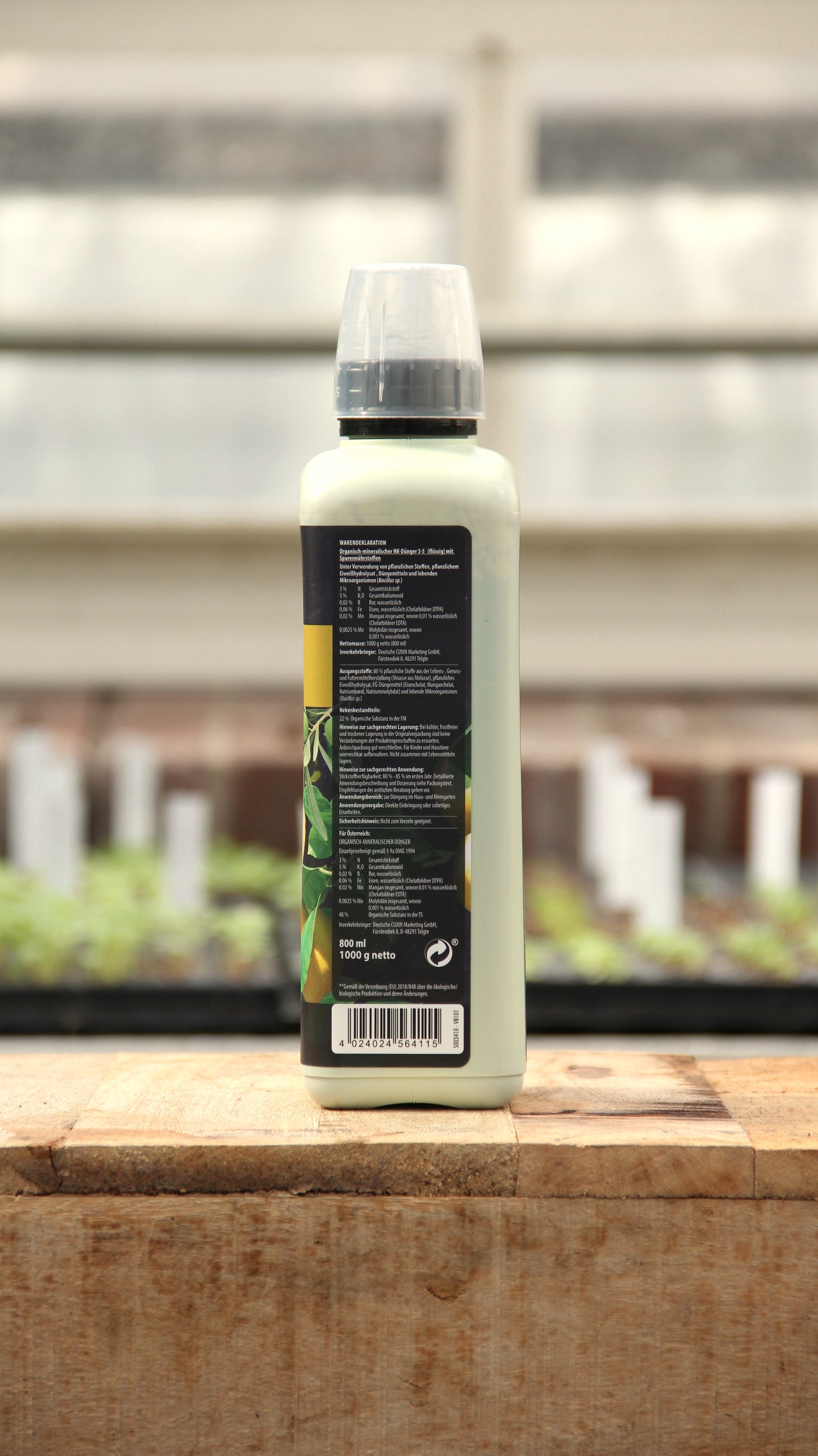 Eine Flasche Cuxin | Flüssigdünger Mediterrane Pflanzen & Zitruspflanzen Bio 800 ml mit schwarzem Etikett und klarem Verschluss steht auf Holz. Das Etikett zeigt Text, grüne Blätter, und im Hintergrund sind unscharfe Pflanzen mit weißen Pflanzenmarkern.