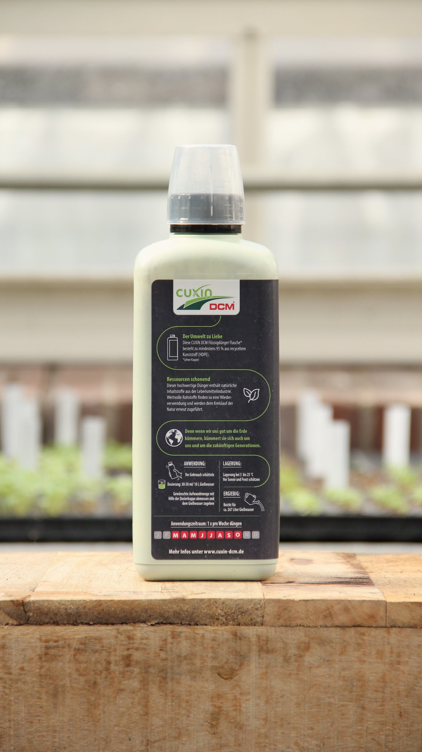 Eine 800-ml-Flasche Cuxin Flüssigdünger Mediterrane Pflanzen & Zitruspflanzen Bio steht auf einer hölzernen Unterlage mit einem Gewächshaus als Hintergrund. Auf dem Etikett sind Produktangaben und Anweisungen zu finden.