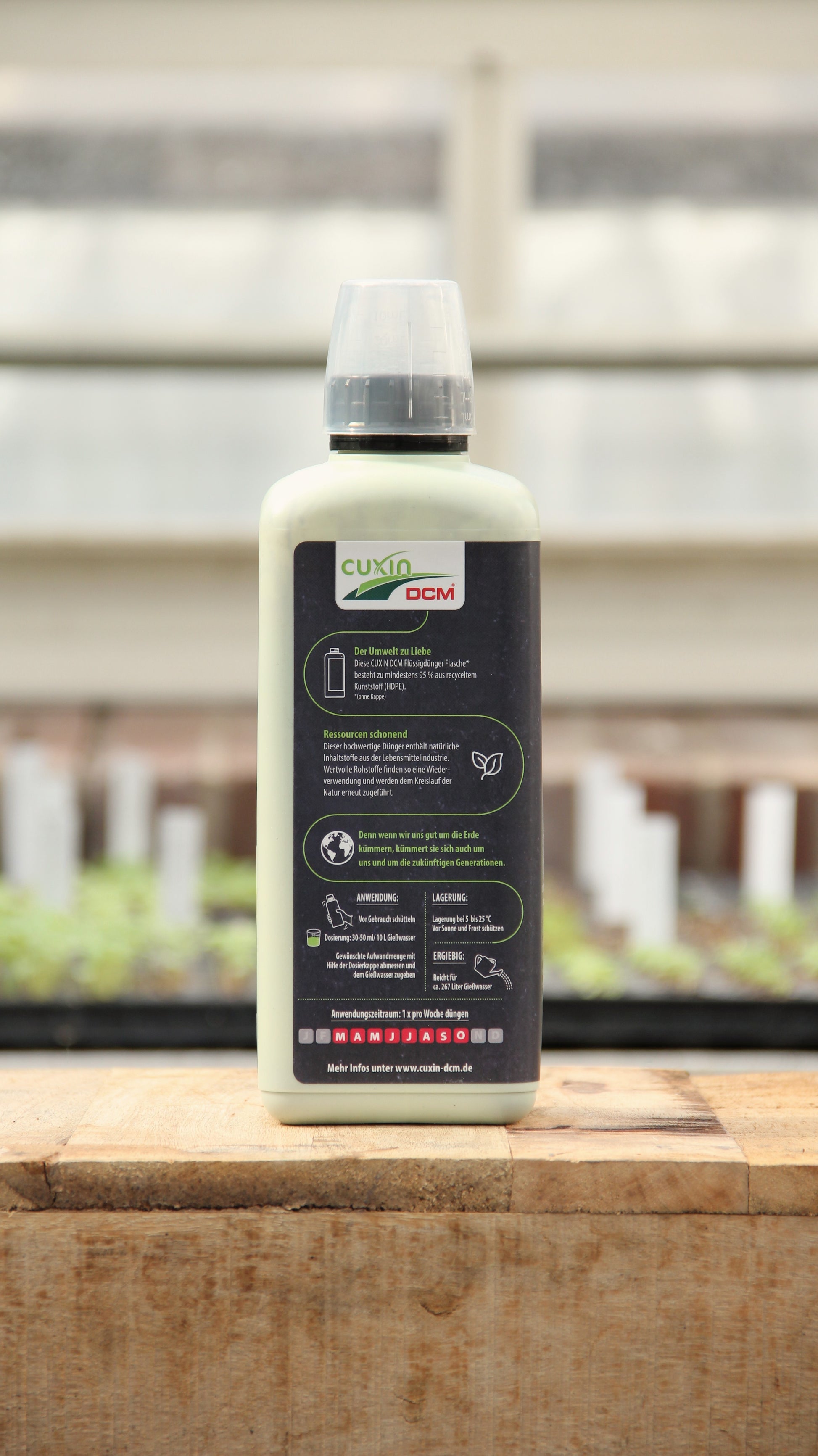 Eine 800-ml-Flasche Cuxin Flüssigdünger Mediterrane Pflanzen & Zitruspflanzen Bio steht auf einer hölzernen Unterlage mit einem Gewächshaus als Hintergrund. Auf dem Etikett sind Produktangaben und Anweisungen zu finden.