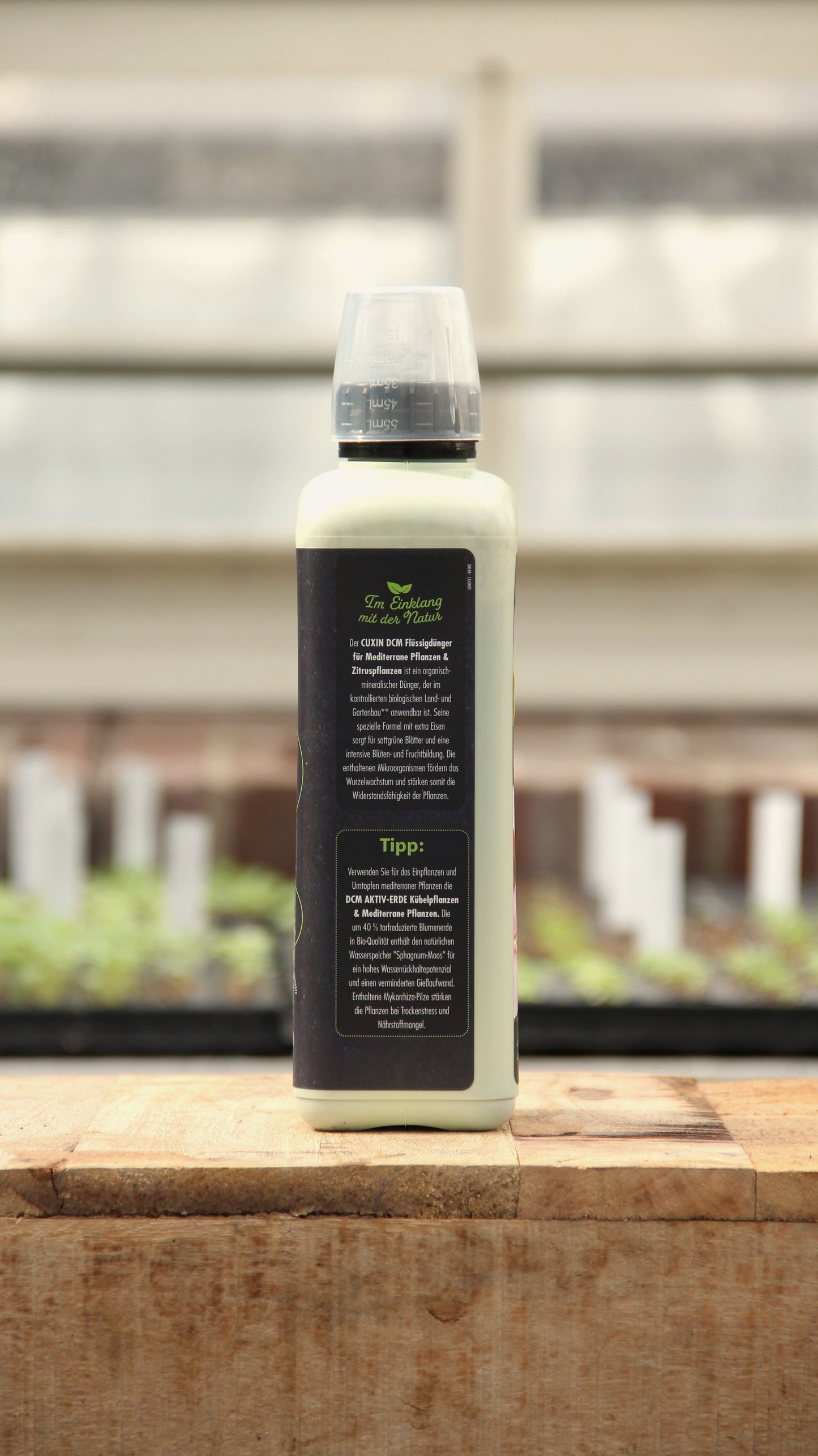 Eine Cuxin Flüssigdünger Mediterrane Pflanzen & Zitruspflanzen Bio 800 ml Flasche mit dunklem deutschem Etikett und durchsichtigem Verschluss steht auf einem hölzernen, verschwommenen Gewächshausregal mit Pflanzen im Hintergrund. Das Etikett ist mit einer grünen Spitze versehen.