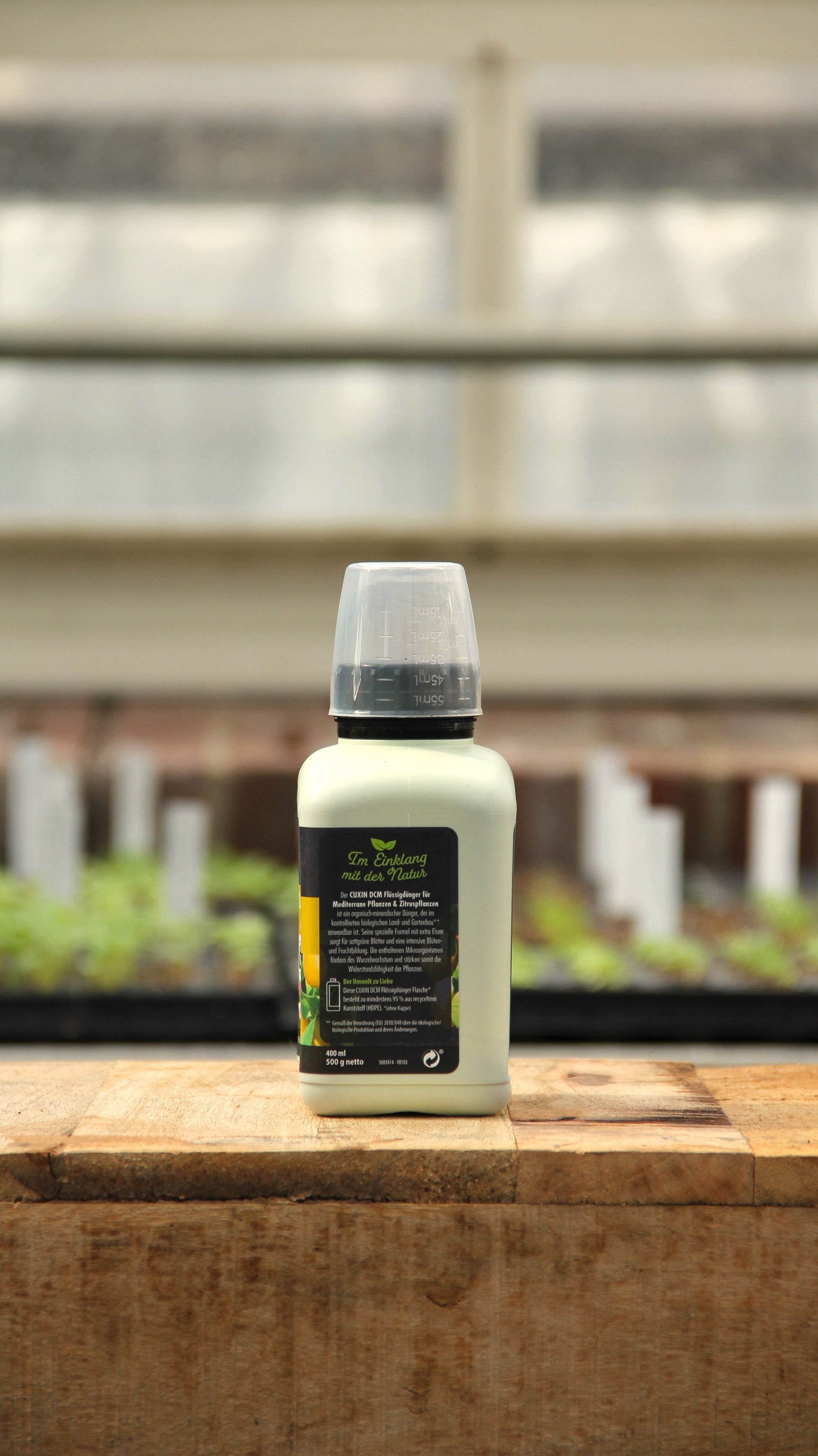 Eine kleine weiße Plastikflasche von Cuxin Flüssigdünger Mediterrane Pflanzen & Zitruspflanzen Bio 400 ml mit grauem Verschluss und grün-gelbem Etikett steht auf einer Holzfläche; im Hintergrund sind unscharfe Pflanzen und weiße Markierungen zu sehen.