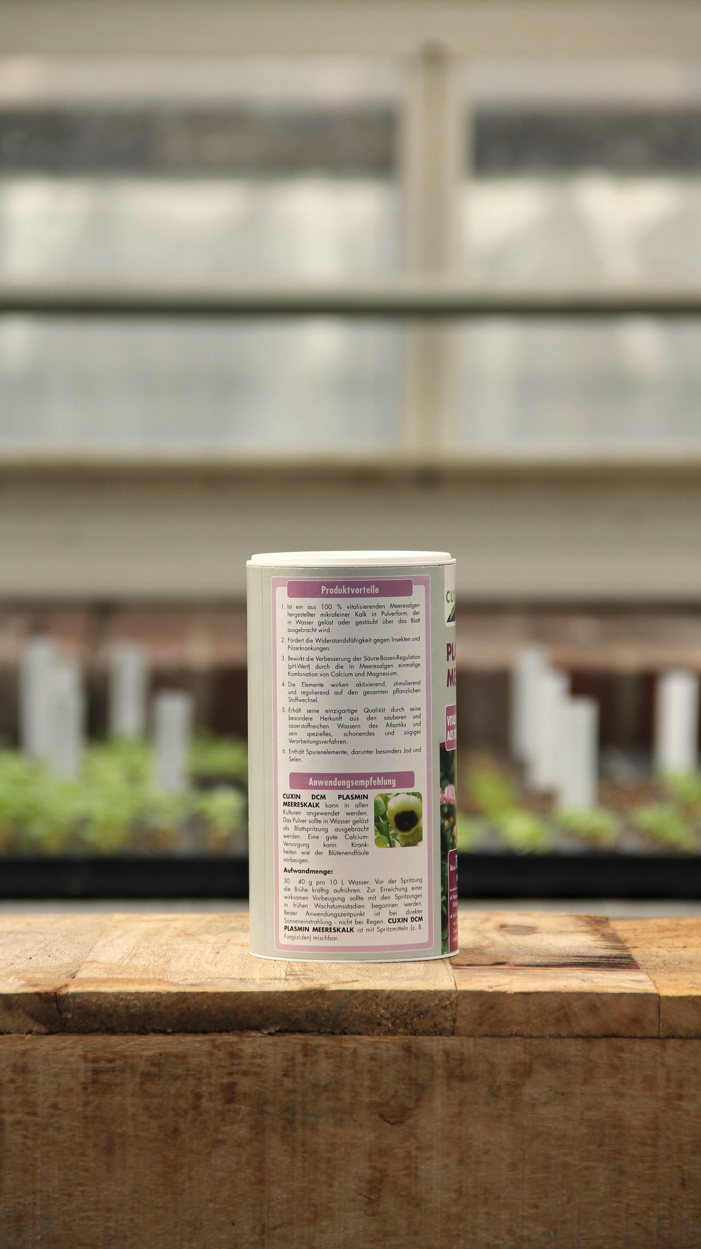 Ein Behälter der Königlichen Gartenakademie Cuxin | Plasmin Meereskalk 250 g steht auf einer hölzernen Unterlage, auf der eine detaillierte Produktinformation zu sehen ist. Im unscharfen Hintergrund sind ein Gewächshaus und Pflanzenmarker zu sehen.