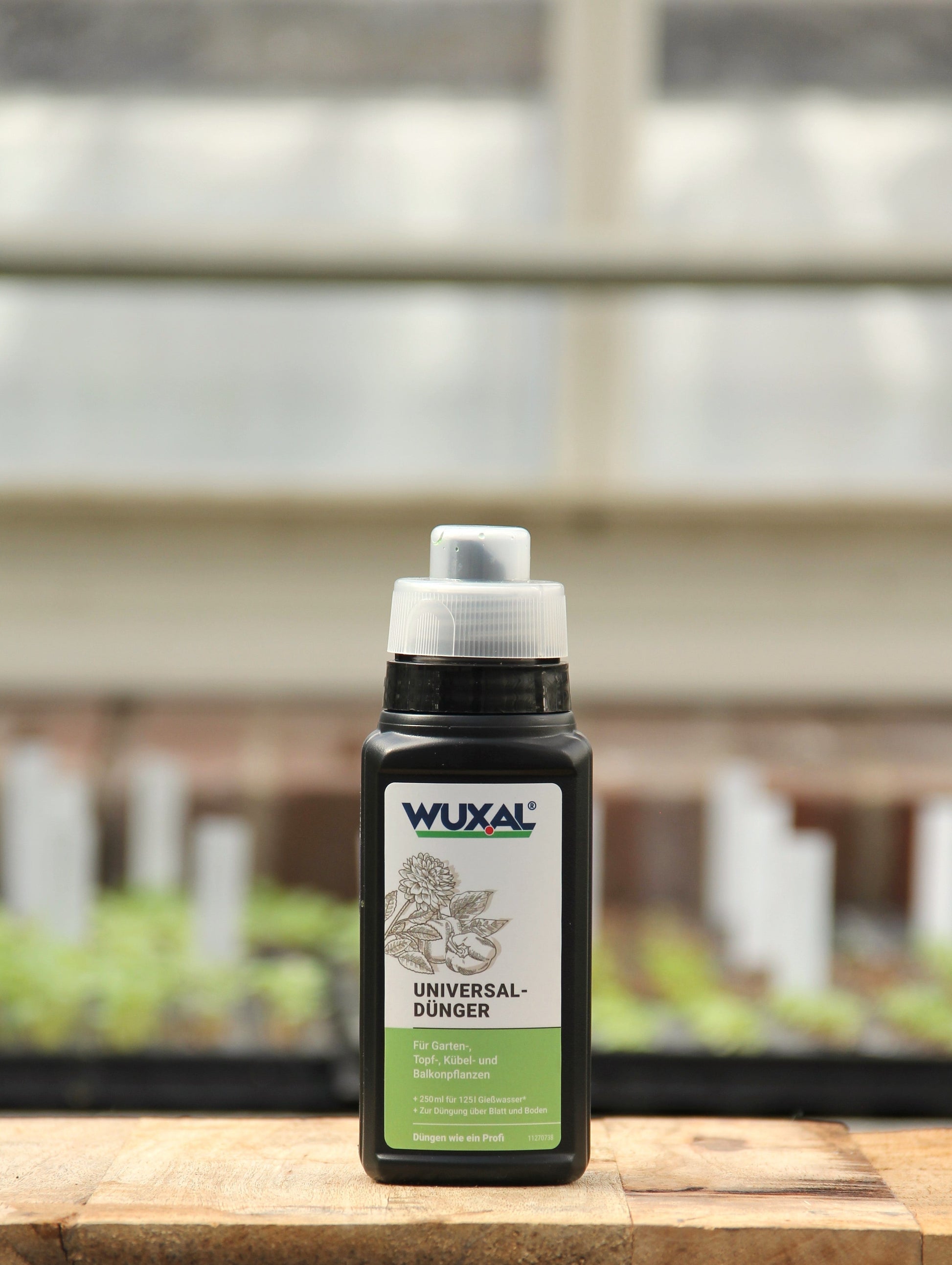 Eine Flasche Wuxal | Universaldünger 250 ml von Wuxal steht auf einer hölzernen Oberfläche, mit unscharfen grünen Pflanzen und weißen Pflanzenetiketten im Hintergrund.