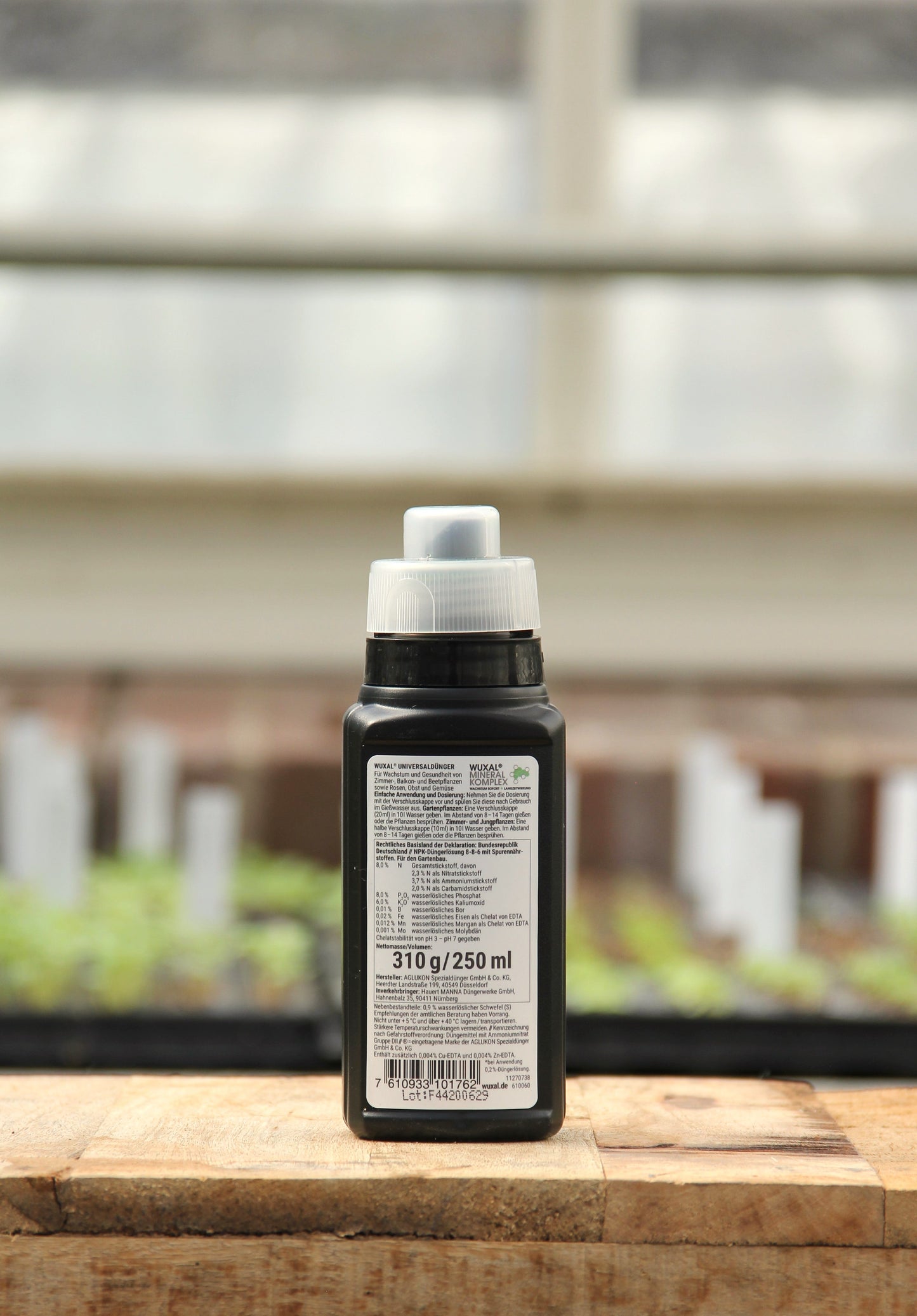 Eine schwarze 250-ml-Plastikflasche von Wuxal Universaldünger mit weißem Verschluss und beschriftetem Text steht auf einer hölzernen Oberfläche in einem Innenraum, vor einem unscharfen Hintergrund aus Pflanzen und weißen Markern.