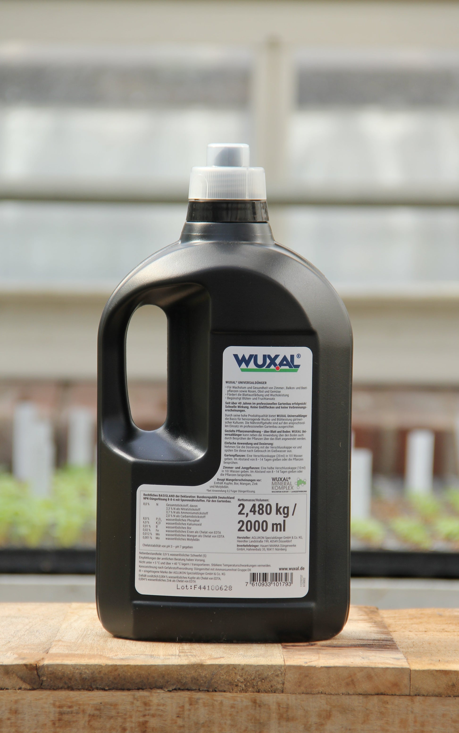 Eine schwarze Wuxal Universaldünger 2 l Kunststoffflasche mit weißem Etikett und Henkel steht auf einer Holzfläche, beschriftet mit 2,480 kg / 2000 ml. Im Hintergrund sind unscharfe Gewächshauspflanzen zu sehen.