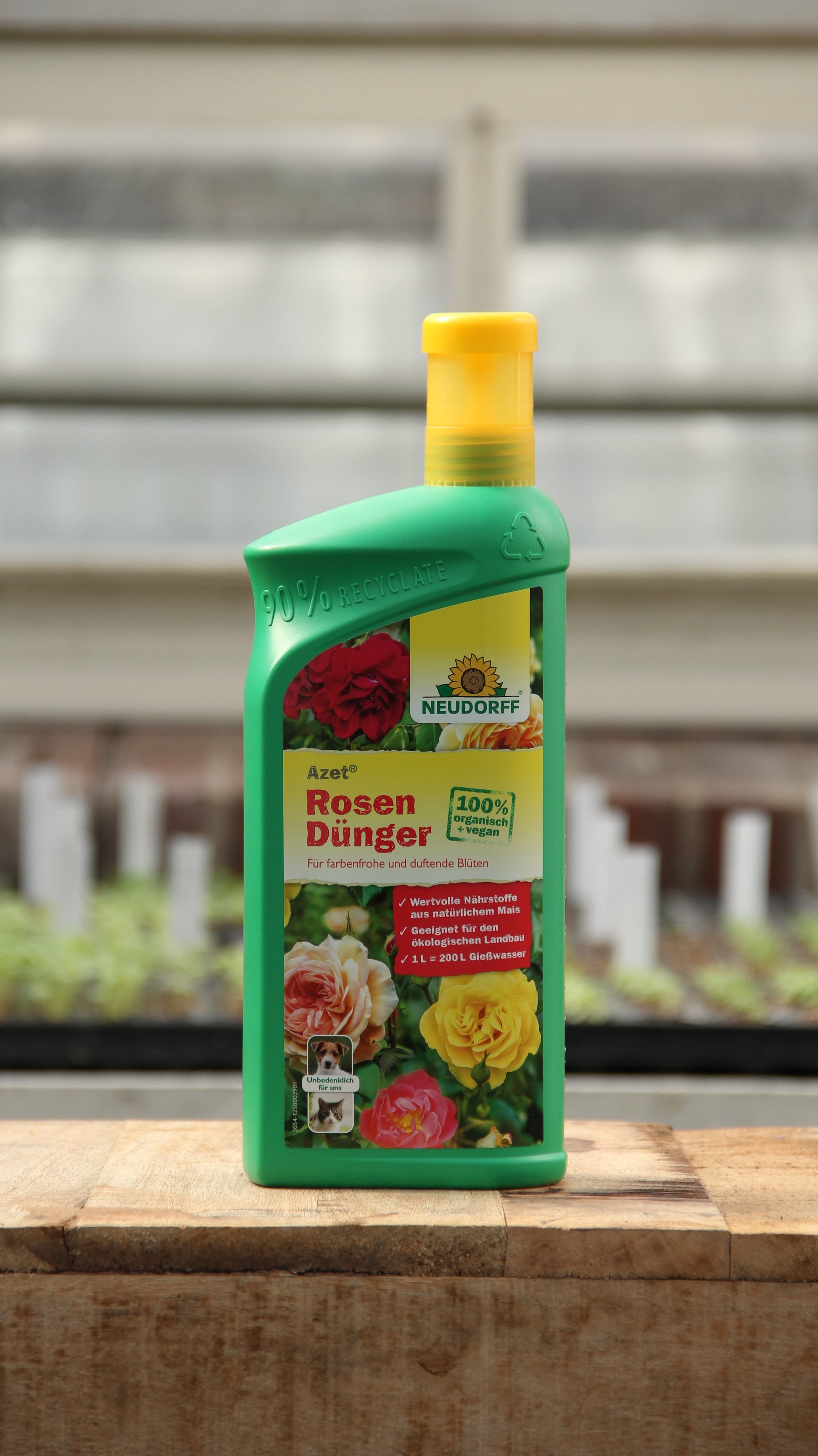 Eine grüne 1-Liter-Plastikflasche von Neudorff Azet Rosendünger flüssig steht auf Holz, perfekt zum Düngen von Rosen, mit einem verschwommenen Gewächshaus im Hintergrund.