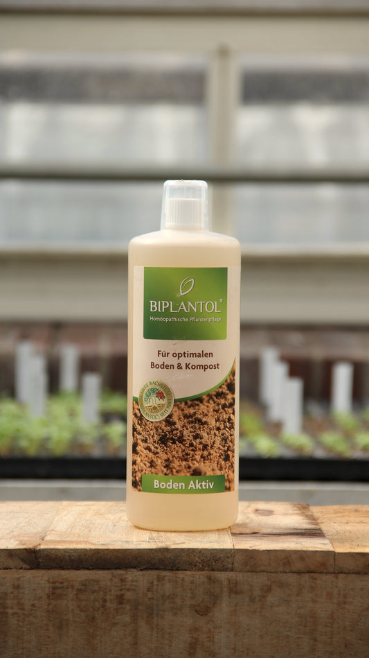 Eine 1-Liter-Flasche Bipantol Biplantol 'Boden Aktiv' Natürlicher Bodenaktivator steht auf einer Holzfläche, im Hintergrund sind Gartenbeete und ein Gewächshaus zu sehen.