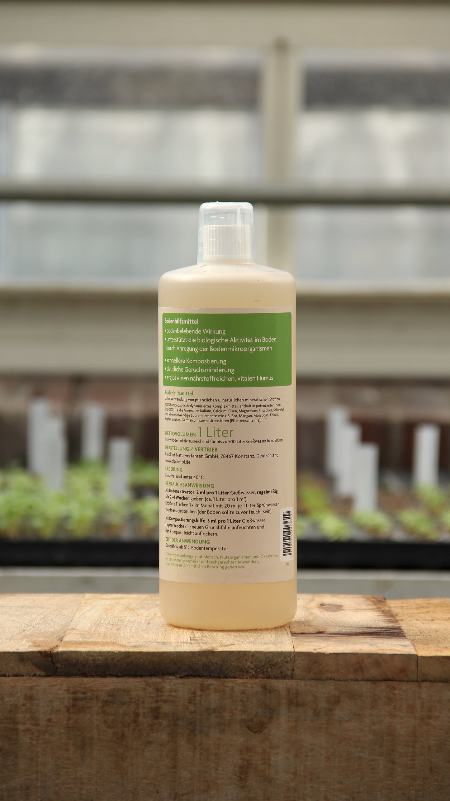 Eine 1-Liter-Flasche Biplantol 'Boden Aktiv' Natürlicher Bodenaktivator von Bipantol mit weißer Kappe und grünem Etikett steht auf Holz; der deutsche Text suggeriert, dass es den Boden verbessert, und im Hintergrund des Gewächshauses sind unscharfe Setzlinge zu sehen.