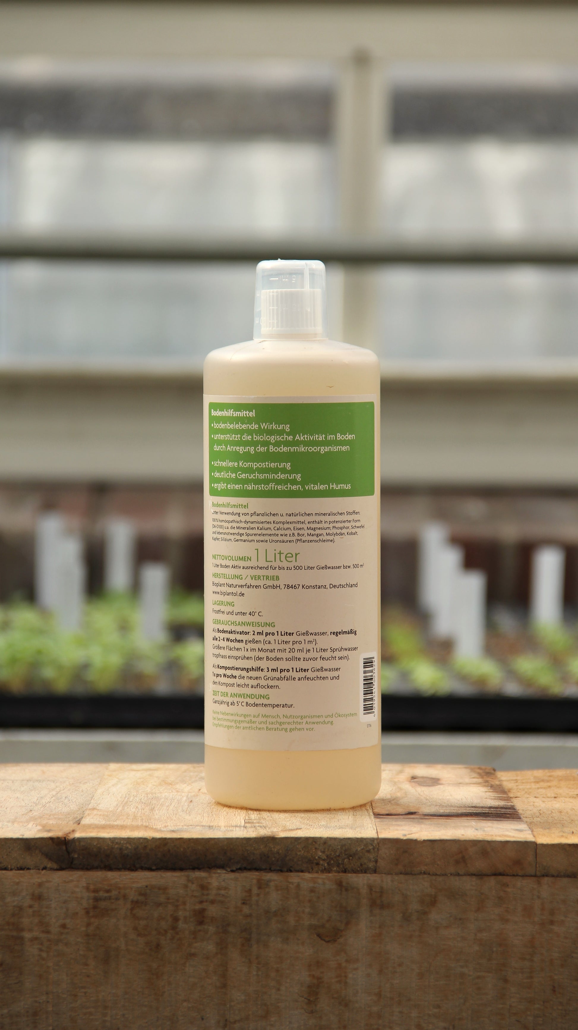 Eine 1-Liter-Flasche Biplantol 'Boden Aktiv' Natürlicher Bodenaktivator von Bipantol mit weißer Kappe und grünem Etikett steht auf Holz; der deutsche Text suggeriert, dass es den Boden verbessert, und im Hintergrund des Gewächshauses sind unscharfe Setzlinge zu sehen.