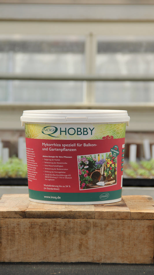 Ein 5-Liter-Plastikbecher mit INOQ Hobby | Mykorrhiza Wurzelkraft für gesunde Pflanzen von Inoq steht auf einer Holzfläche, im Hintergrund sind ein Gewächshaus und Topfpflanzen zu sehen.