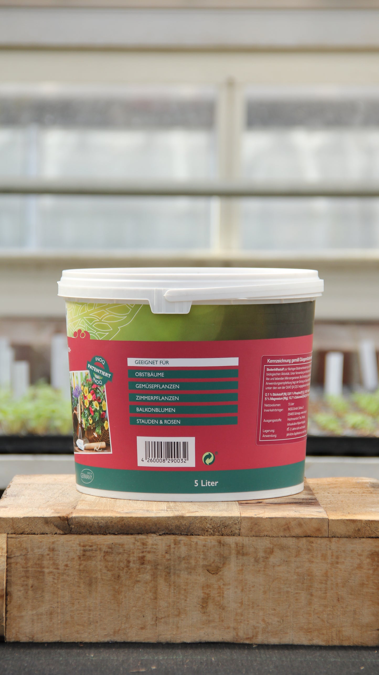Ein 5-Liter-Behälter von INOQ Hobby mit Mykorrhiza Wurzelkraft für gesunde Pflanzen steht auf einer Holzfläche in einem Gewächshaus, sein buntes Inoq-Etikett mit deutscher Anleitung hebt sich von einem sanft verschwommenen Hintergrund ab.