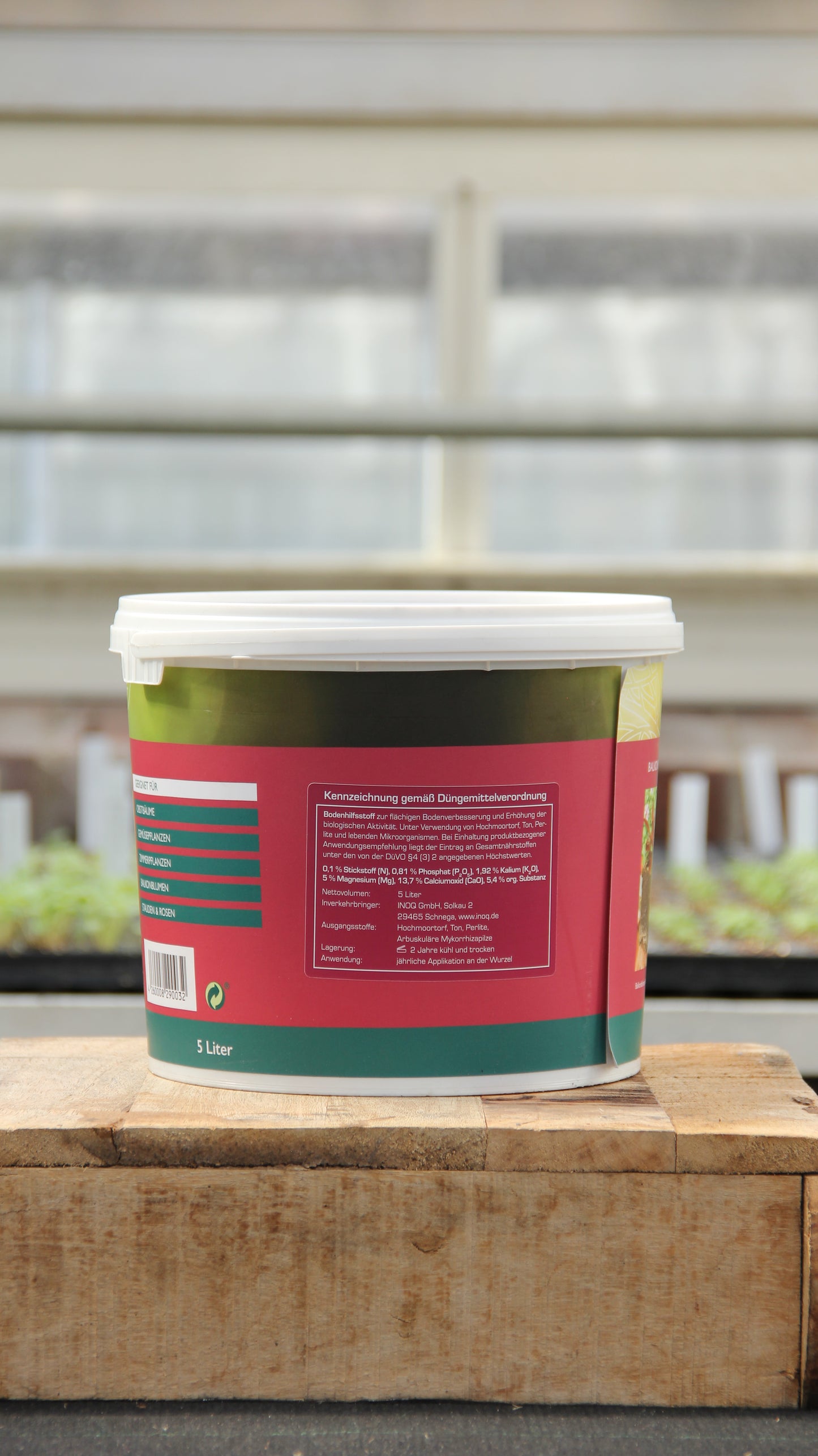 Ein 5-Liter-Behälter von INOQ Hobby Mykorrhiza Wurzelkraft für gesunde Pflanzen mit weißem Deckel und rot-grünem Etikett steht auf Holz, umgeben von grünen Pflanzen und verschwommenen Gewächshausfenstern im Hintergrund.