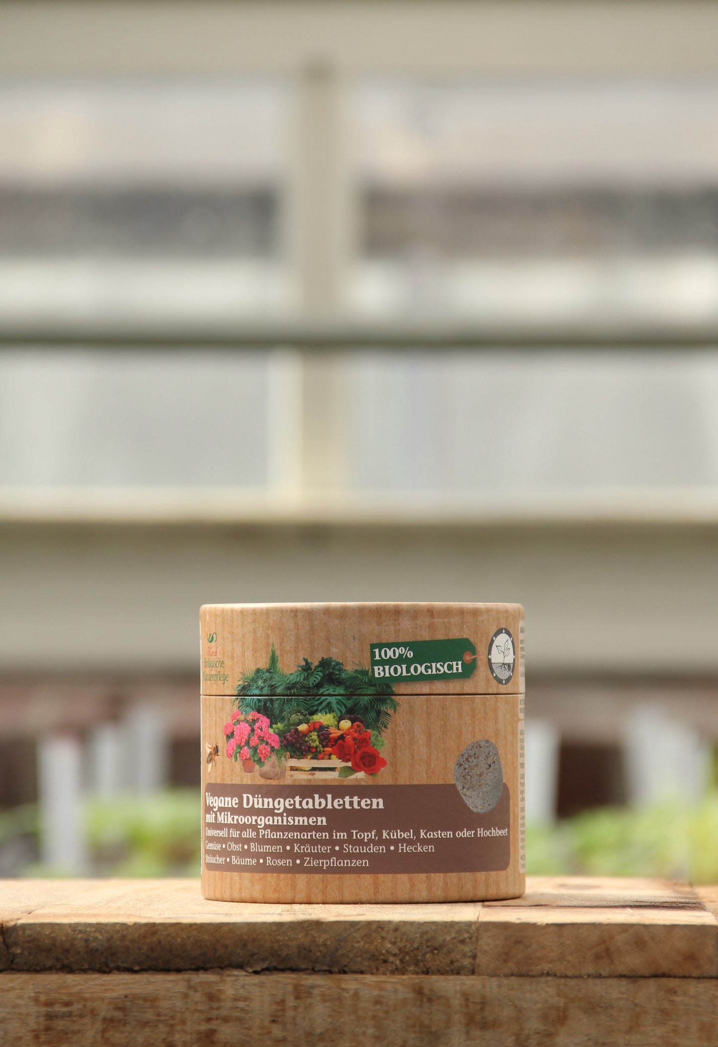 Ein zylindrischer Behälter mit AMN | Vegane Düngertabletten 60 Stk. von AMN mit einem Etikett, das Tomaten und grüne Blätter zeigt, steht auf einer Holzfläche vor einem unscharfen Hintergrund.