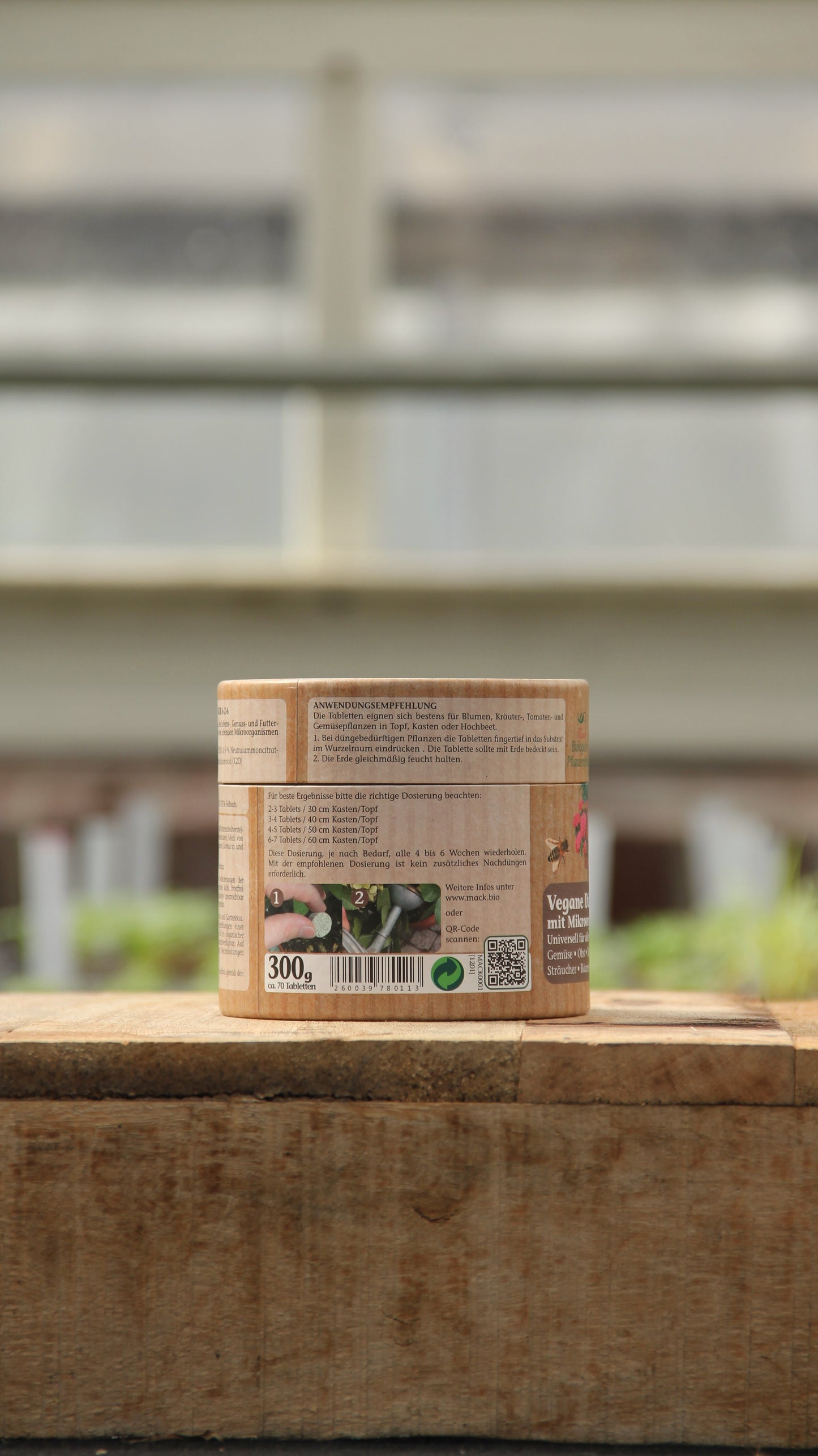 Ein kleiner zylindrischer Behälter mit der Aufschrift "AMN | Vegane Düngertabletten 60 Stk." von AMN steht auf einer hölzernen Fläche im Freien. Das beigefarbene Etikett mit deutschem Text ist vor einem verschwommenen Hintergrund aus Grünpflanzen und Fenstern zu sehen.