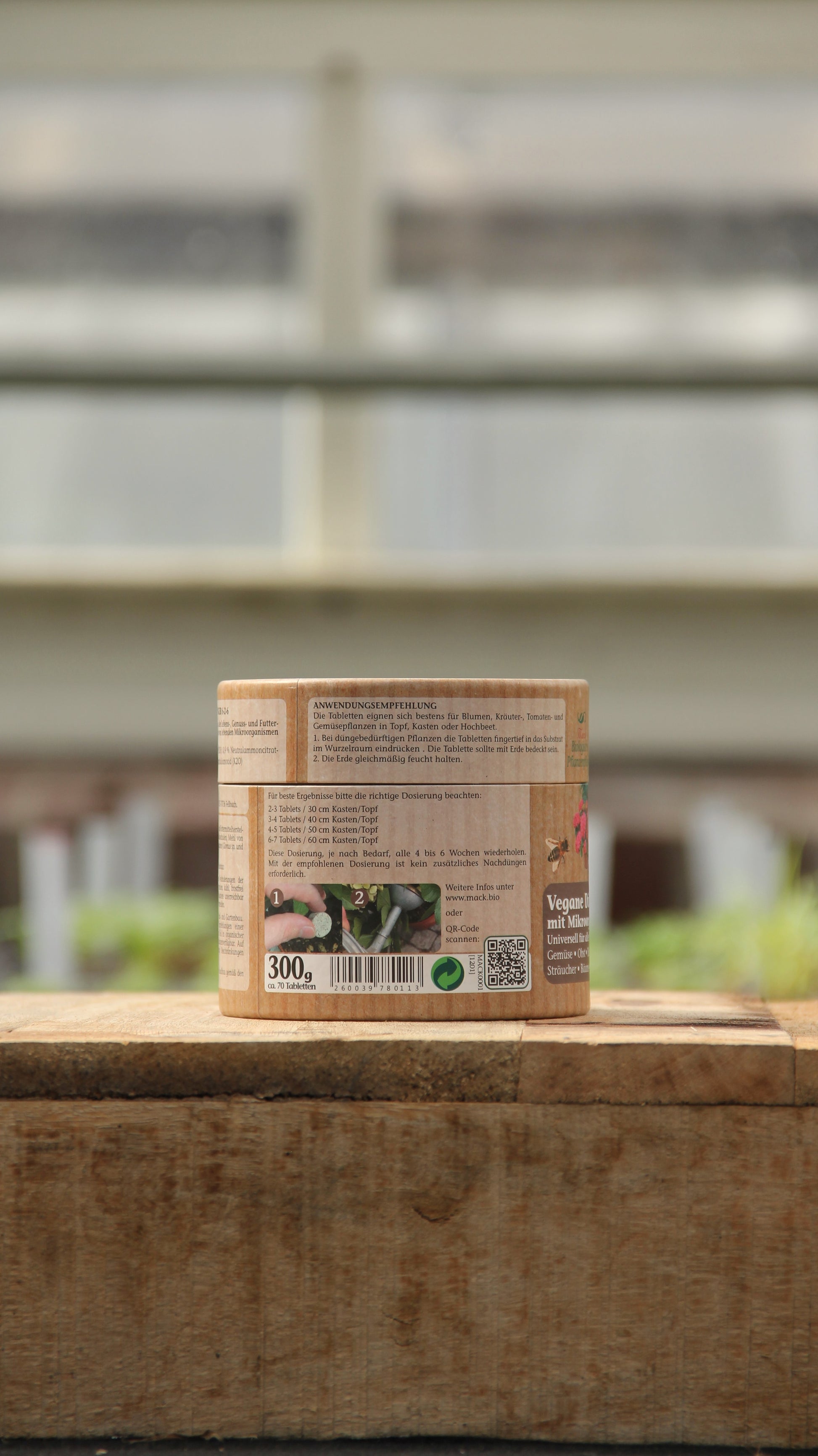 Ein kleiner zylindrischer Behälter mit der Aufschrift "AMN | Vegane Düngertabletten 60 Stk." von AMN steht auf einer hölzernen Fläche im Freien. Das beigefarbene Etikett mit deutschem Text ist vor einem verschwommenen Hintergrund aus Grünpflanzen und Fenstern zu sehen.