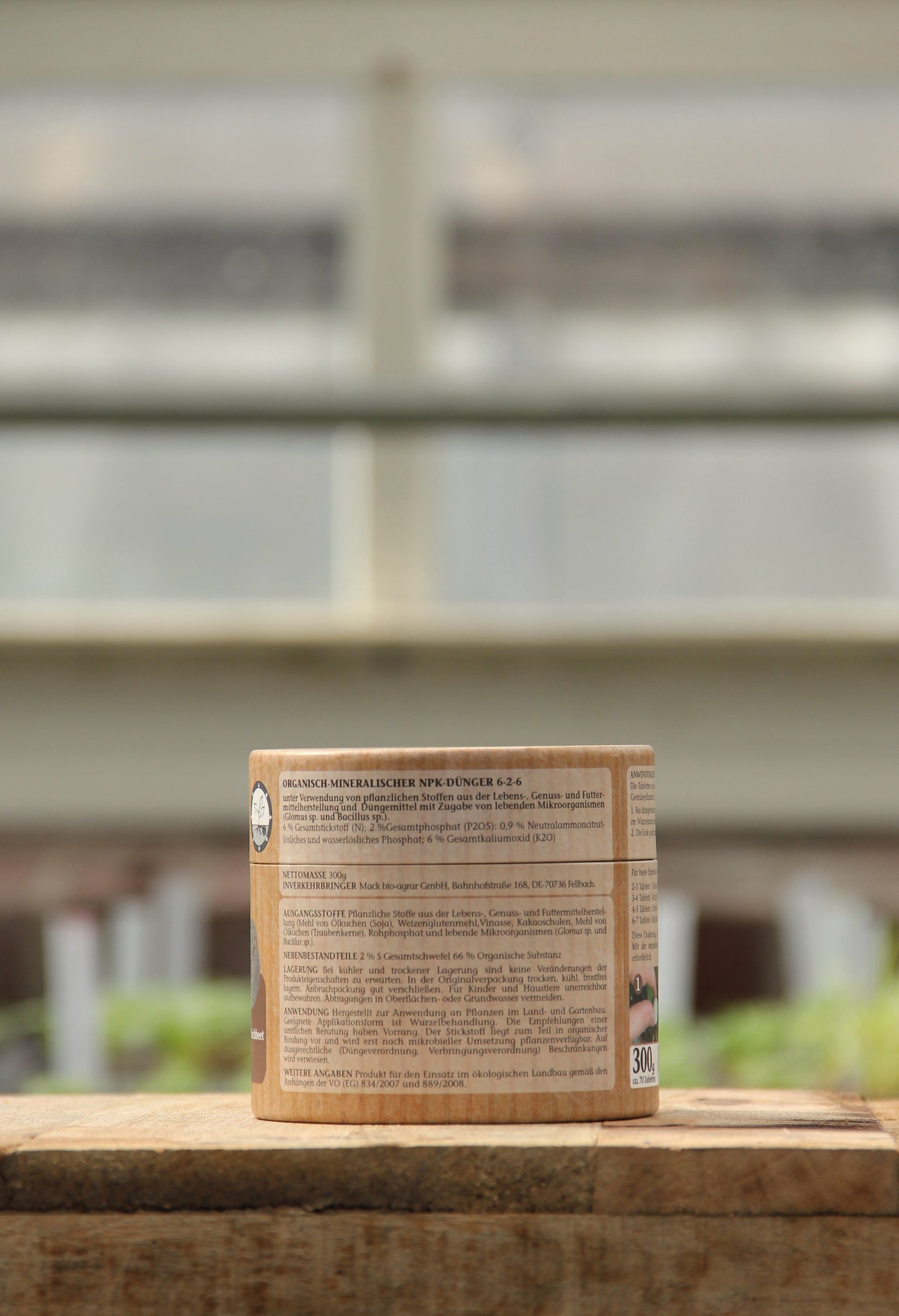 Ein brauner zylindrischer AMN-Behälter mit der Aufschrift "Vegane Düngertabletten 60 Stk." steht auf einer Holzfläche, hinter der sich verschwommene Fenster und Grünpflanzen befinden.