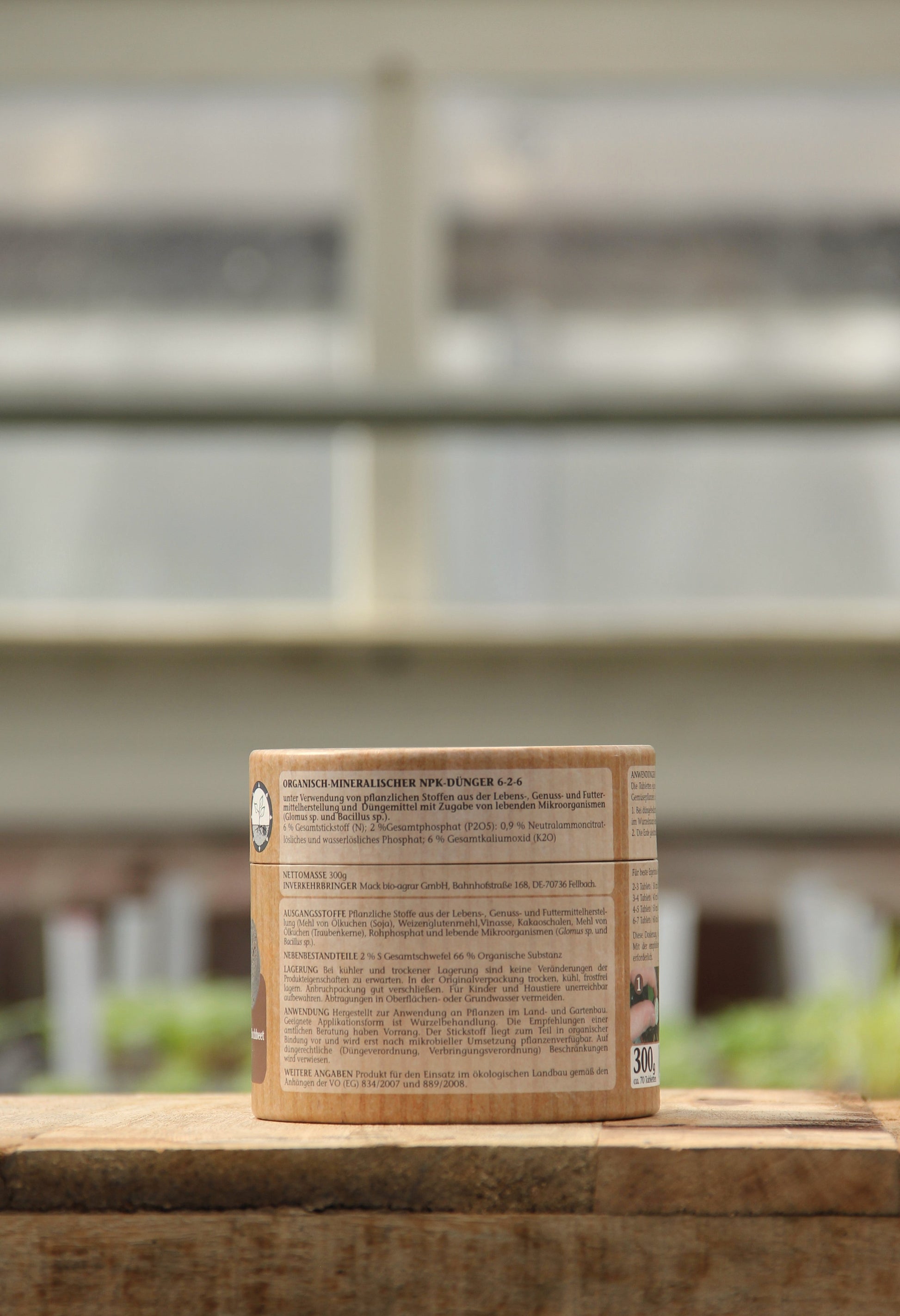 Ein brauner zylindrischer AMN-Behälter mit der Aufschrift "Vegane Düngertabletten 60 Stk." steht auf einer Holzfläche, hinter der sich verschwommene Fenster und Grünpflanzen befinden.